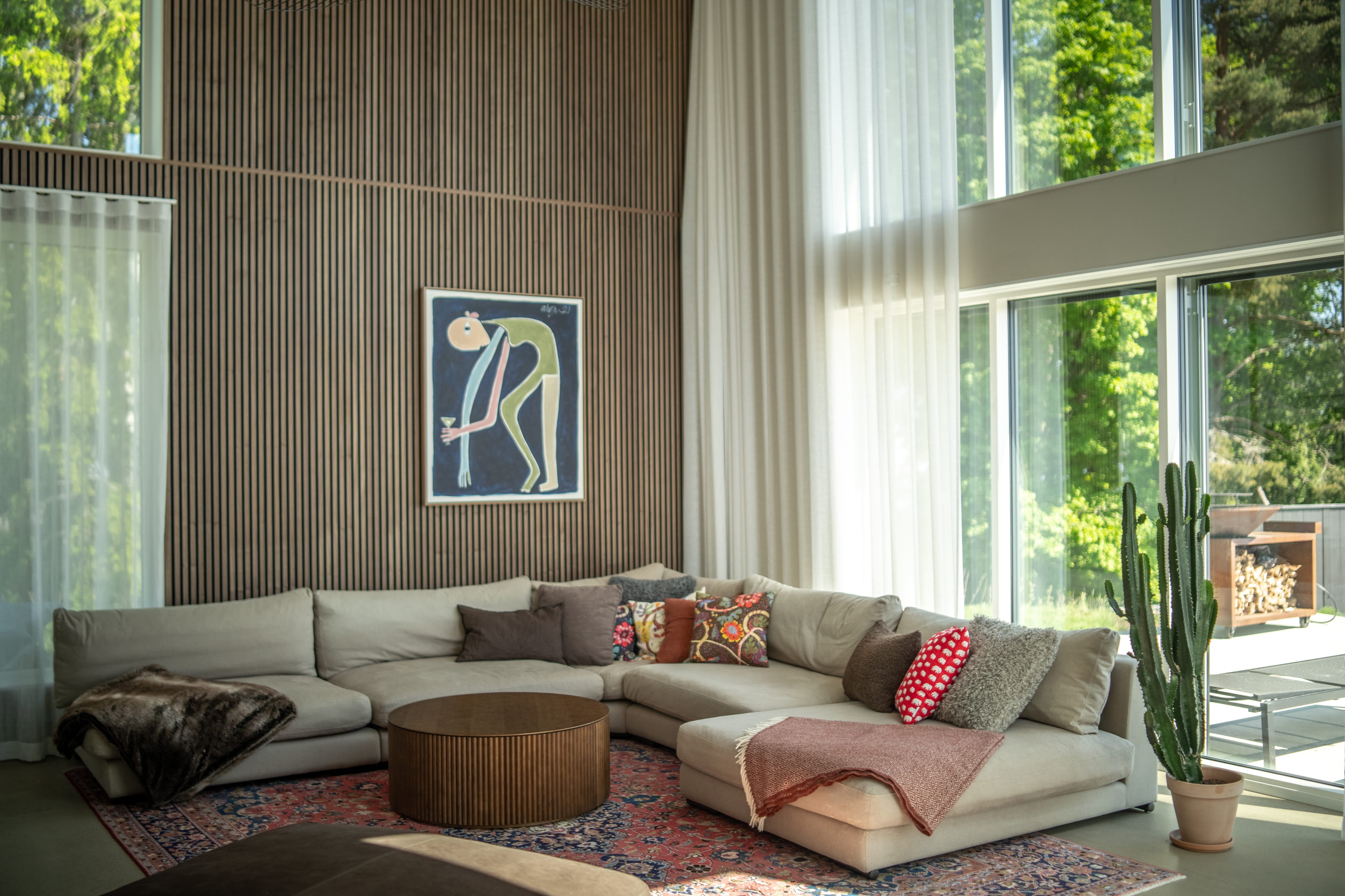Salon moderne avec un grand canapé sectionnel beige, des coussins colorés, une table basse ronde, un cactus et des fenêtres allant du sol au plafond avec des rideaux transparents et des panneaux acoustiques nordiques.