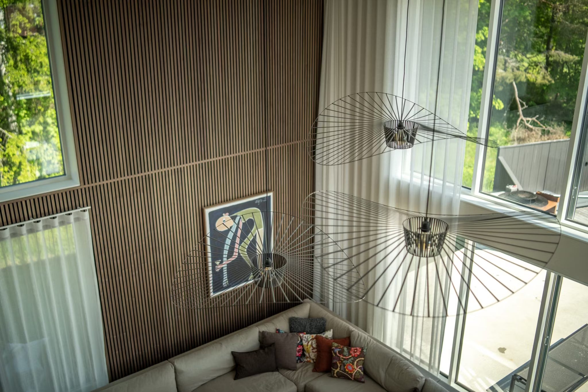 Salon moderne avec des murs en bois nordique Akupanel, de grandes fenêtres, des panneaux acoustiques abstraits, un canapé sectionnel et des luminaires minimalistes en fil de fer, vus den haut.