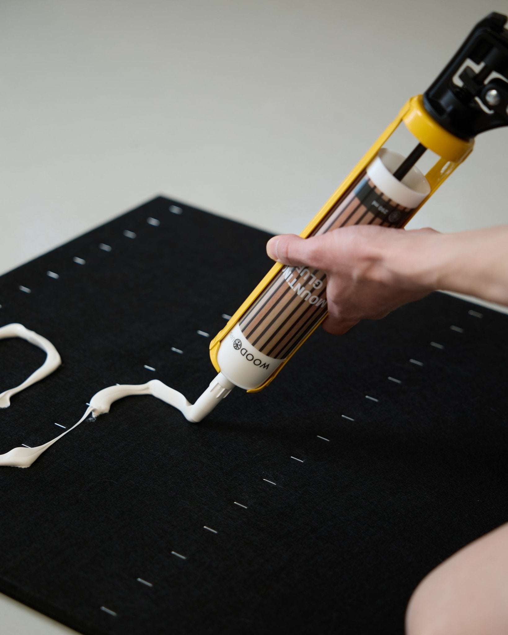 Une main applique de la colle de montage blanche en zigzag avec le pistolet à colle WoodUpp (290 ml) sur une surface black Panneau acoustique.