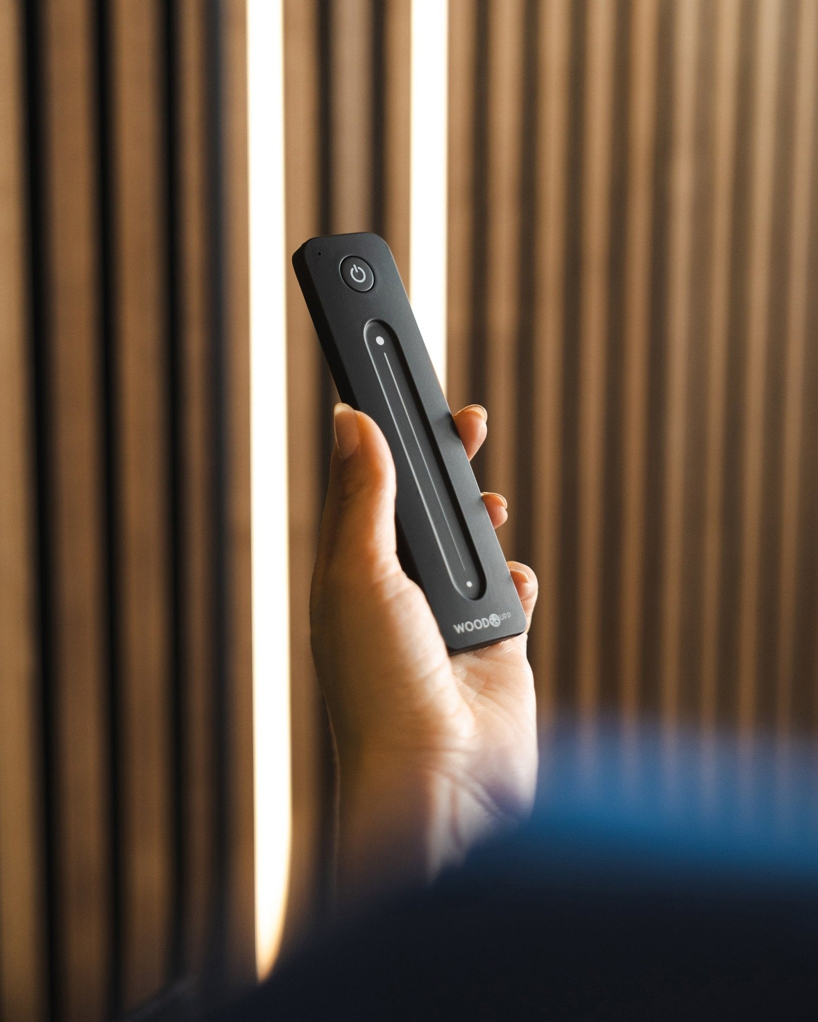 Une main tient une télécommande pour ruban LED WoodUpp black Lumi avec bouton marche/arrêt, placée devant des panneaux acoustiques à lattes WoodUpp ; compatible Philips Hue.