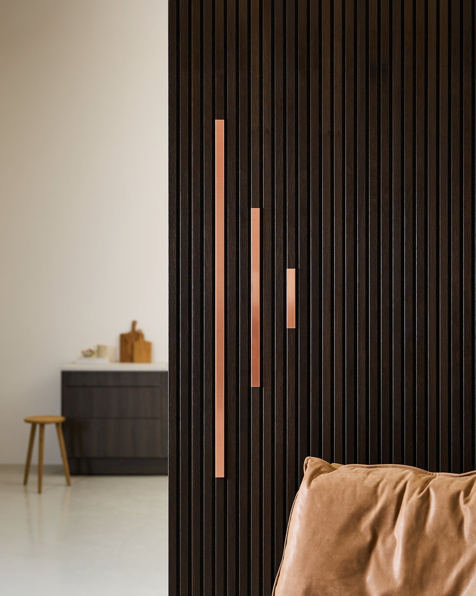 Espace minimaliste avec une cloison acoustique à lattes en bois foncé, le cache WoodUpp Liva en copper, des panneaux de 60 cm avec poignées copper, un fauteuil en cuir fauve et un tabouret d’appoint.