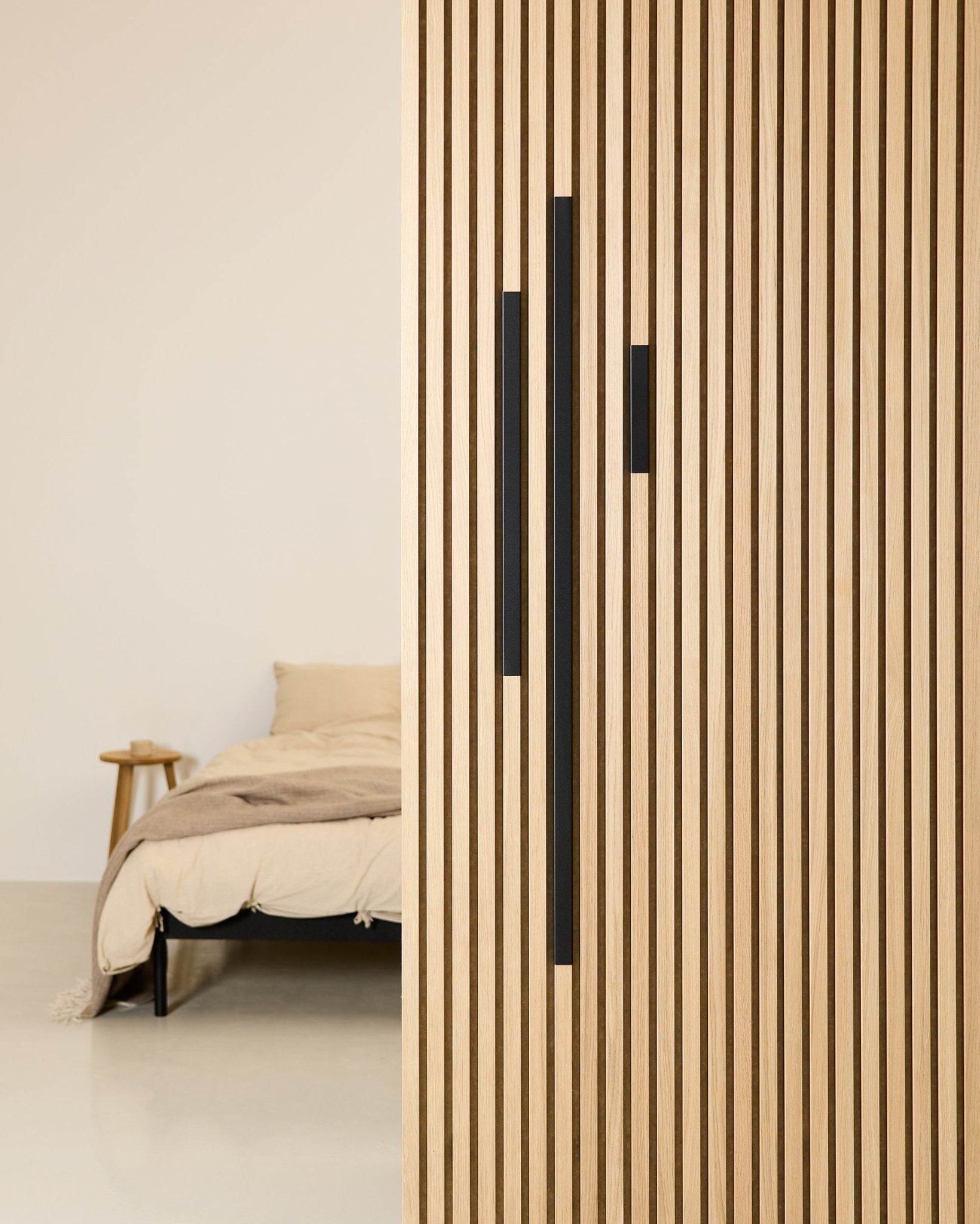 Chambre minimaliste avec un lit et une couverture beige, partiellement dissimulés derrière une cloison WoodUpp Akupanel de 60 cm en aluminium Black, de la série Liva Cover.