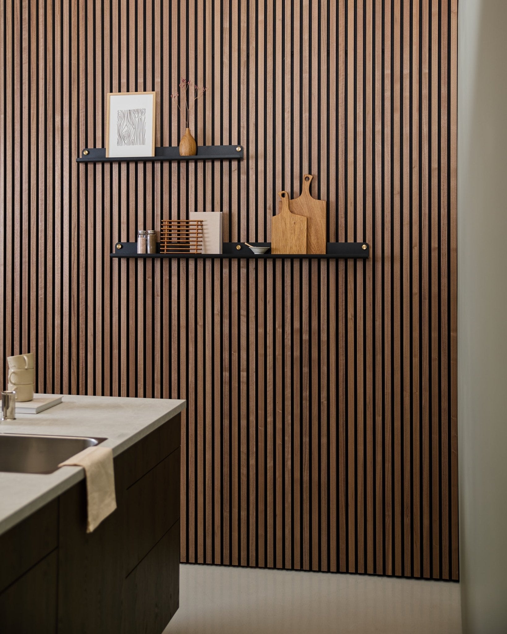 Cuisine moderne avec un panneau acoustique à lattes de style nordique, une étagère étroite WoodUpp Norr de black (115,9 cm) pour la décoration, et une serviette posée sur l’évier.