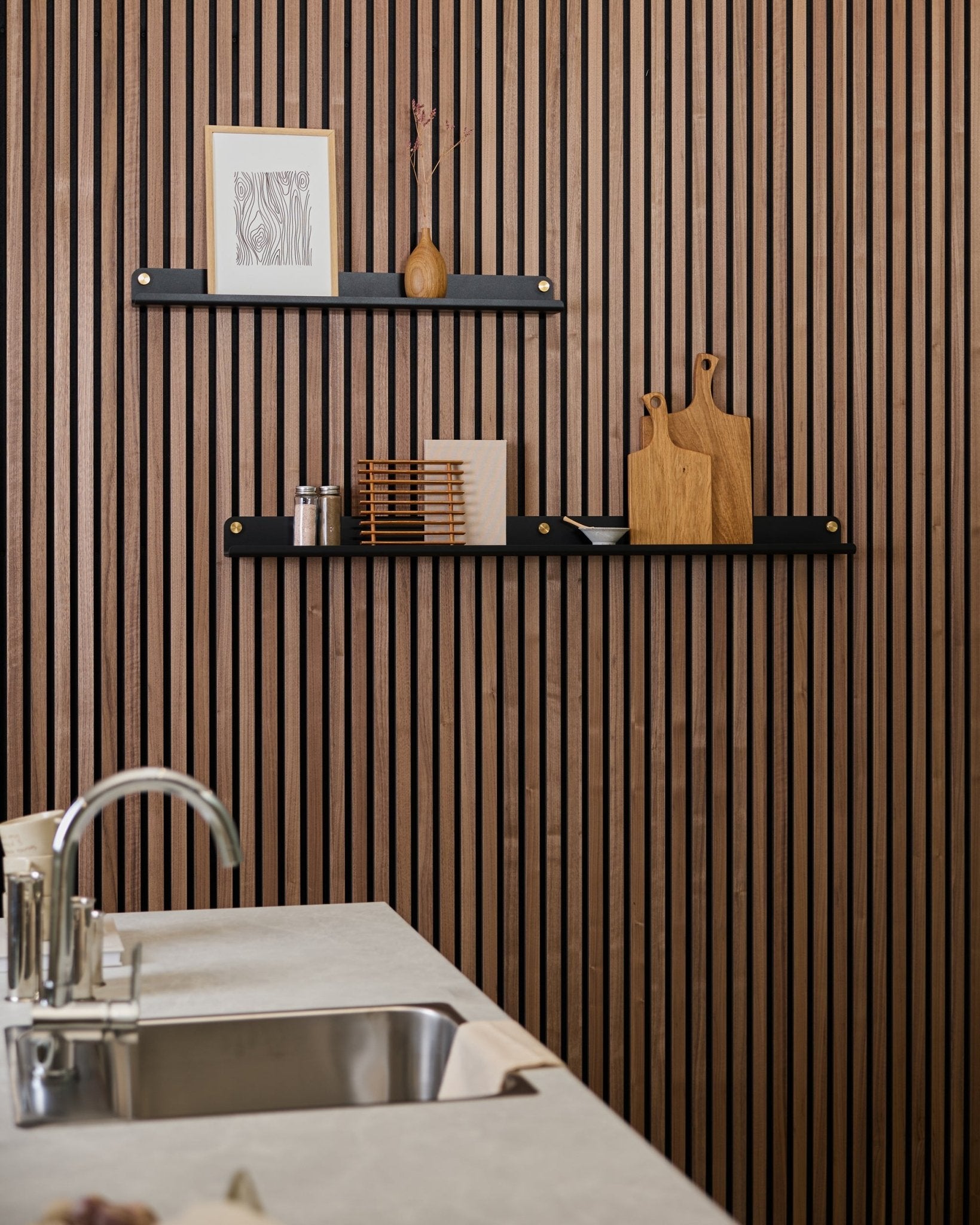 Cuisine minimaliste avec un mur acoustique à lames de bois vertical Akupanel, des étagères black, des planches à découper, des objets déco small et un plan de travail avec évier moderne à l’avant.