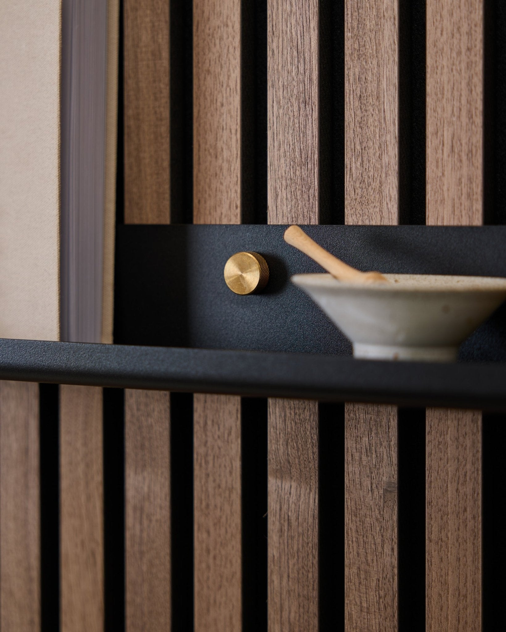 Gros plan sur une étagère étroite WoodUpp Norr de black (115,9 cm), montée sur un panneau acoustique nordique à lattes avec des vis moletées brass, un bouton doré et un bol en céramique avec cuillère.