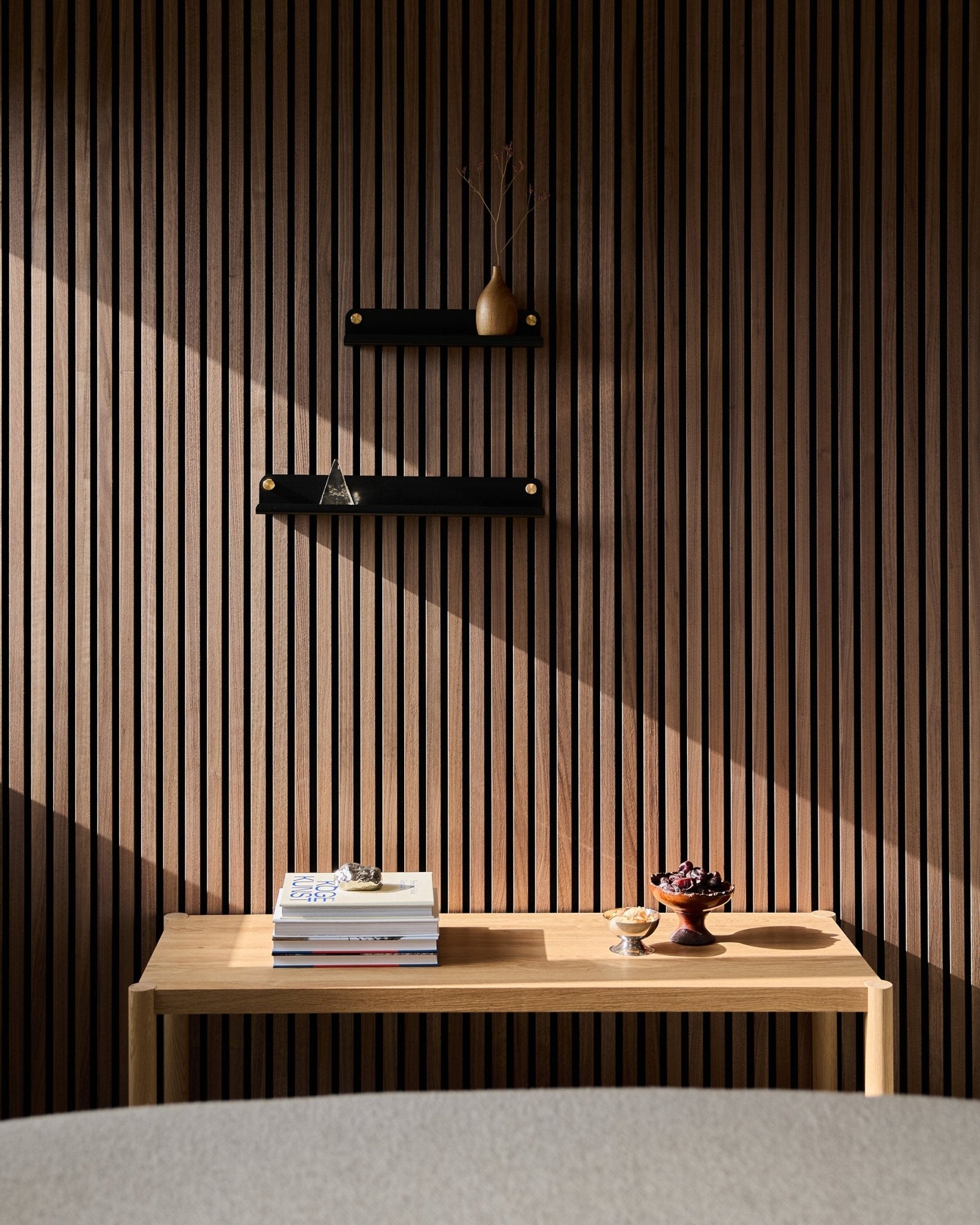Une table en bois minimaliste avec des livres et des bols est placée près d’un panneau à lattes nordique Panneau acoustique, mettant en valeur l’étagère étroite WoodUpp Norr en black (51,9 cm), avec des ombres baignées de soleil au-dessus.