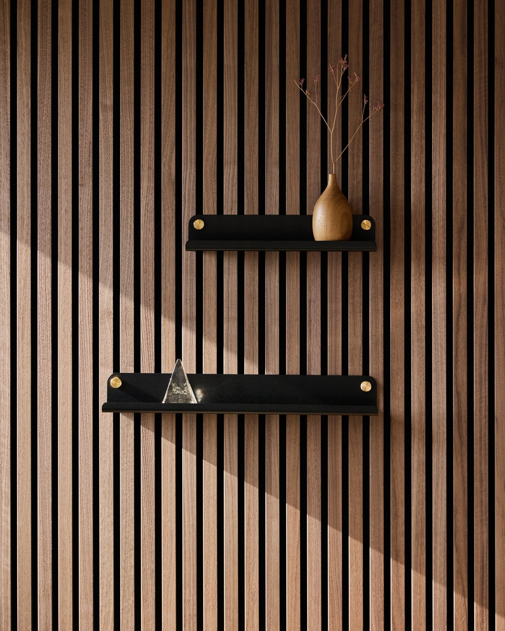 Deux étagères étroites WoodUpp black Norr (35,9 cm) fixées sur un panneau acoustique à lattes de style nordique ; en haut, un vase avec des branches séchées, en bas, une pyramide en verre.
