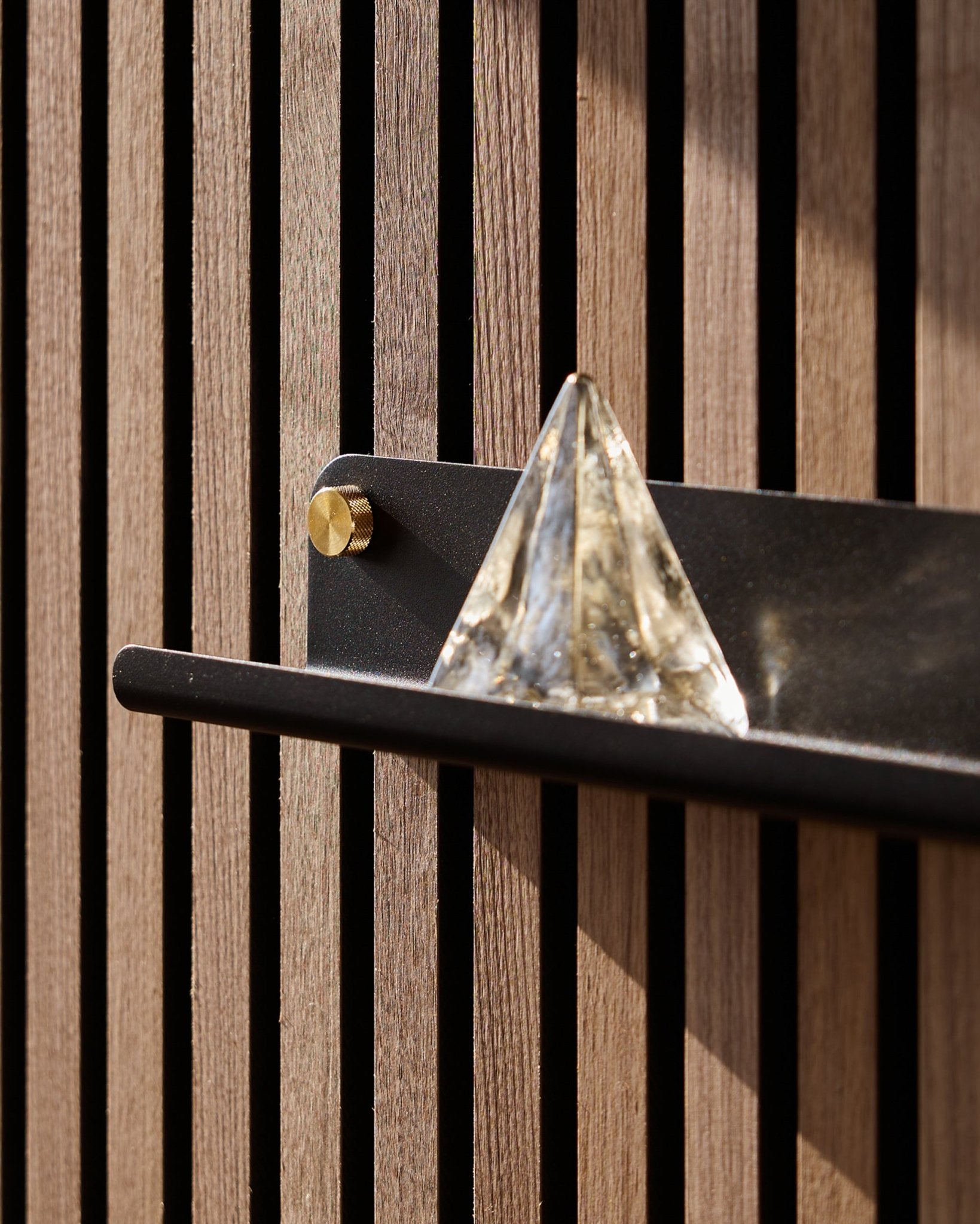 Une pyramide en verre repose sur l’étagère étroite WoodUpp Norr en black (51,9 cm), montée sur un vertical Panneau acoustique et mise en lumière par le soleil.