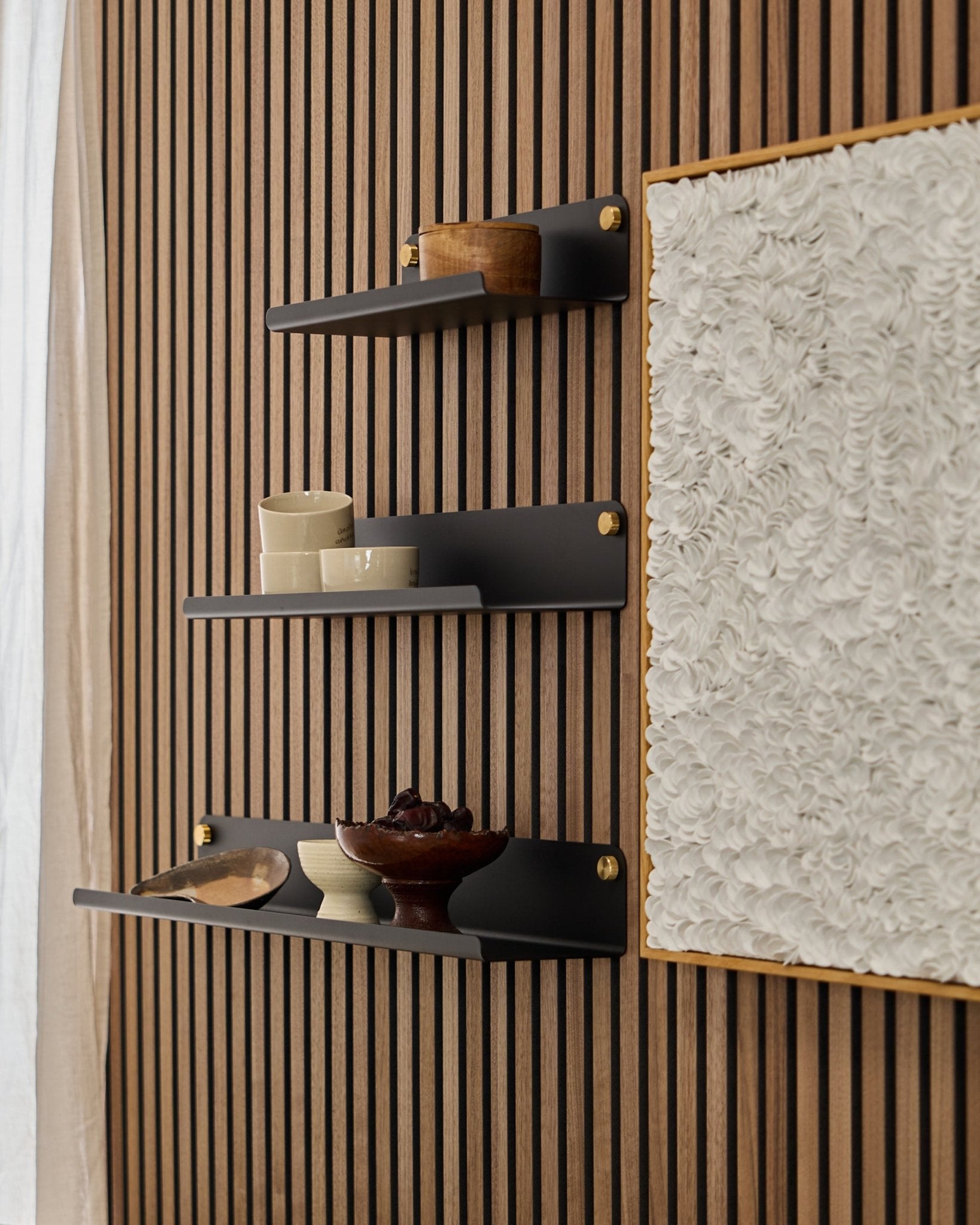 Trois étagères larges WoodUpp Norr black (51,9 cm) montées sur un panneau acoustique en bois WoodUpp Akupanel, à côté d’une œuvre d’art blanche texturée, avec des bols et des contenants en bois.