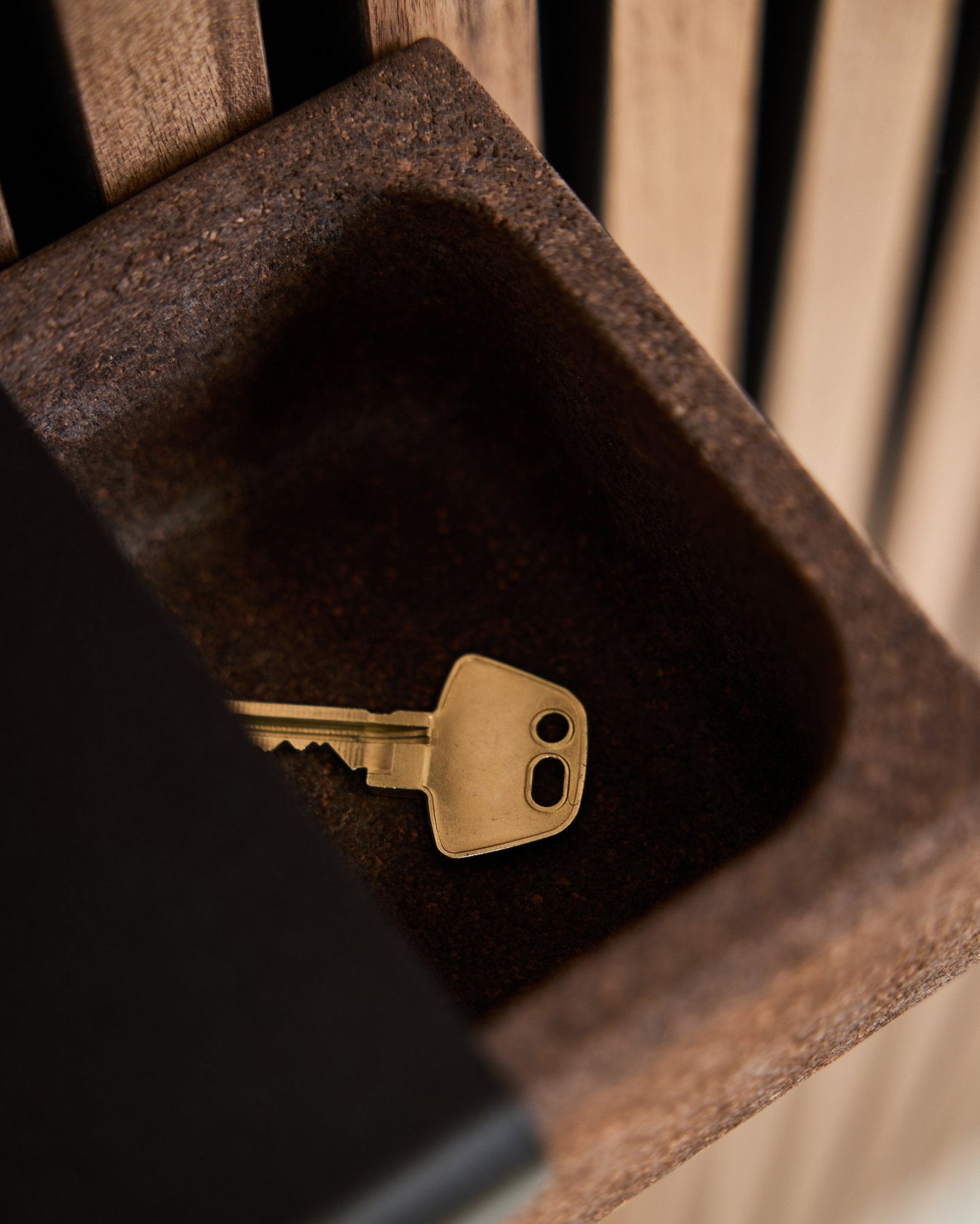 Une clé brass repose dans un Norr Key Tray/Smoked Cork, 35,9 cm, par WoodUpp, posé sur un contenant brown devant un fond de panneau acoustique en bois Akupanel.