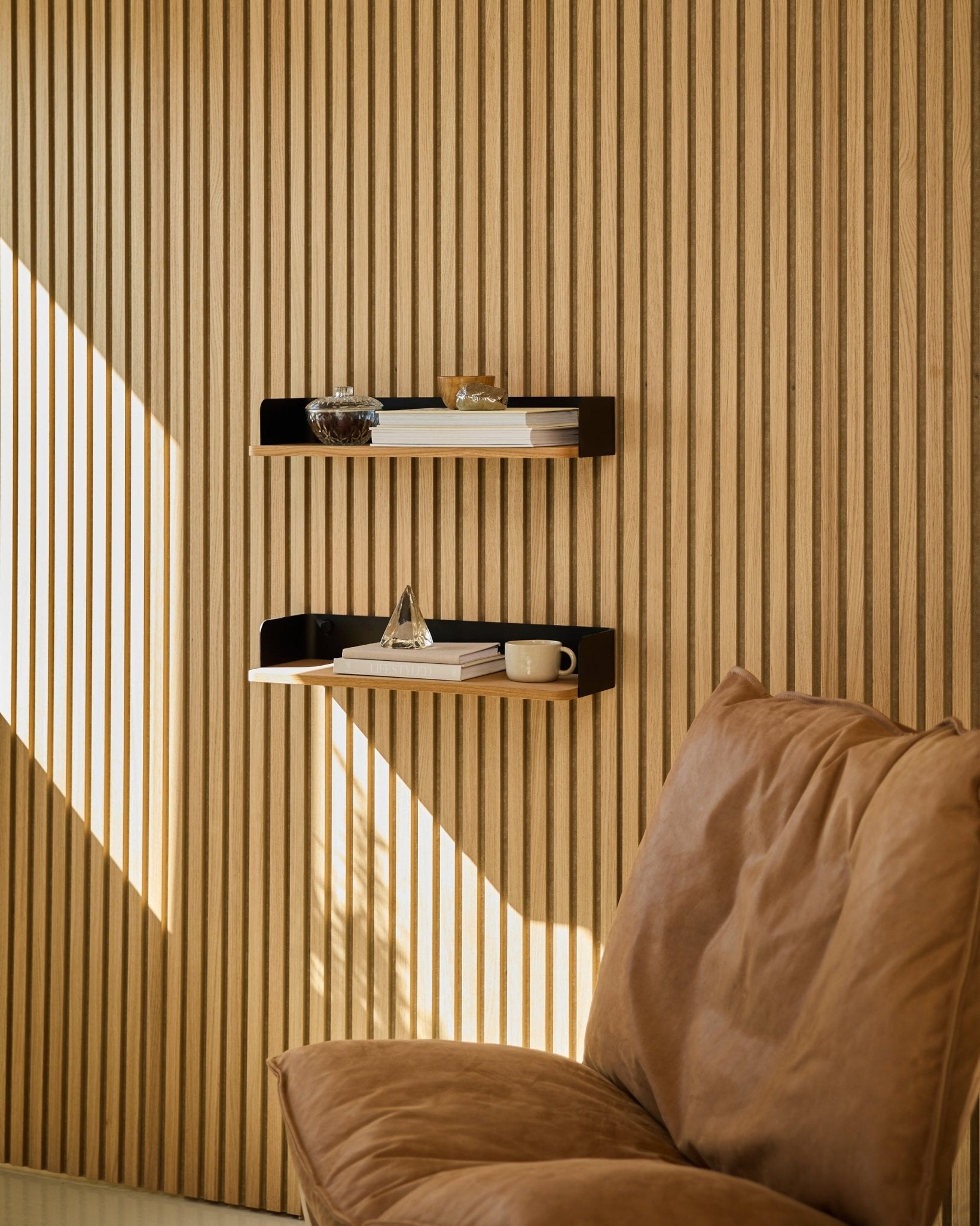Chaise Brown à côté de la bibliothèque WoodUpp Linn en Natural Oak, avec des livres et des objets déco disposés à la lumière naturelle devant un mur en lattes de bois nordique Panneau acoustique.