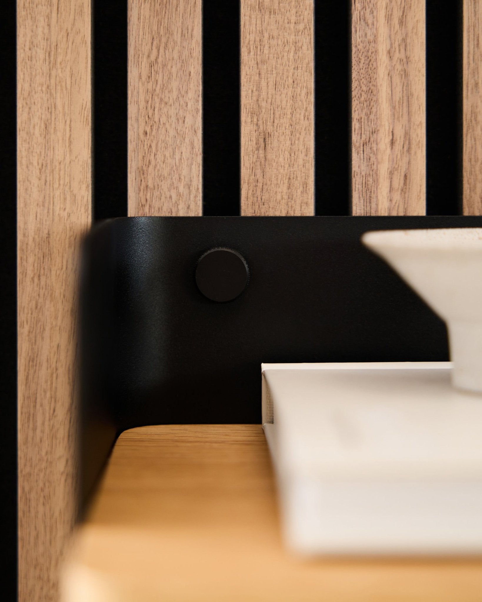 Gros plan sur un panneau acoustique à lattes de bois WoodUpp Akupanel, avec des vis Finger black fixant un support black, à côté de livres blancs et d’un bol.