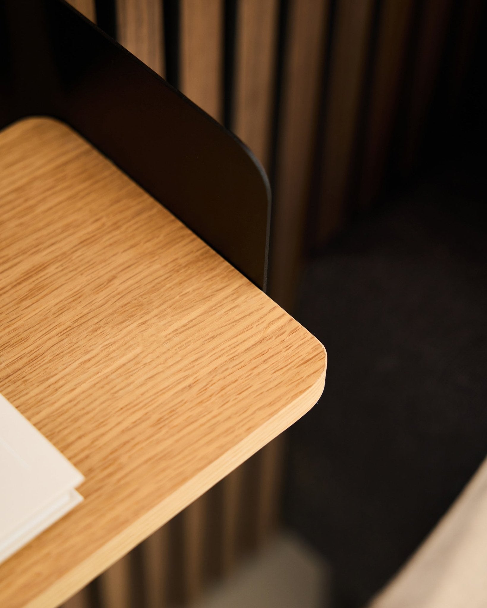Gros plan sur un coin de bureau avec un revêtement mural acoustique en bois black Akupanel, des feuilles blanches, et la bibliothèque WoodUpp Linn en Natural Oak (39,9 cm) en arrière-plan.