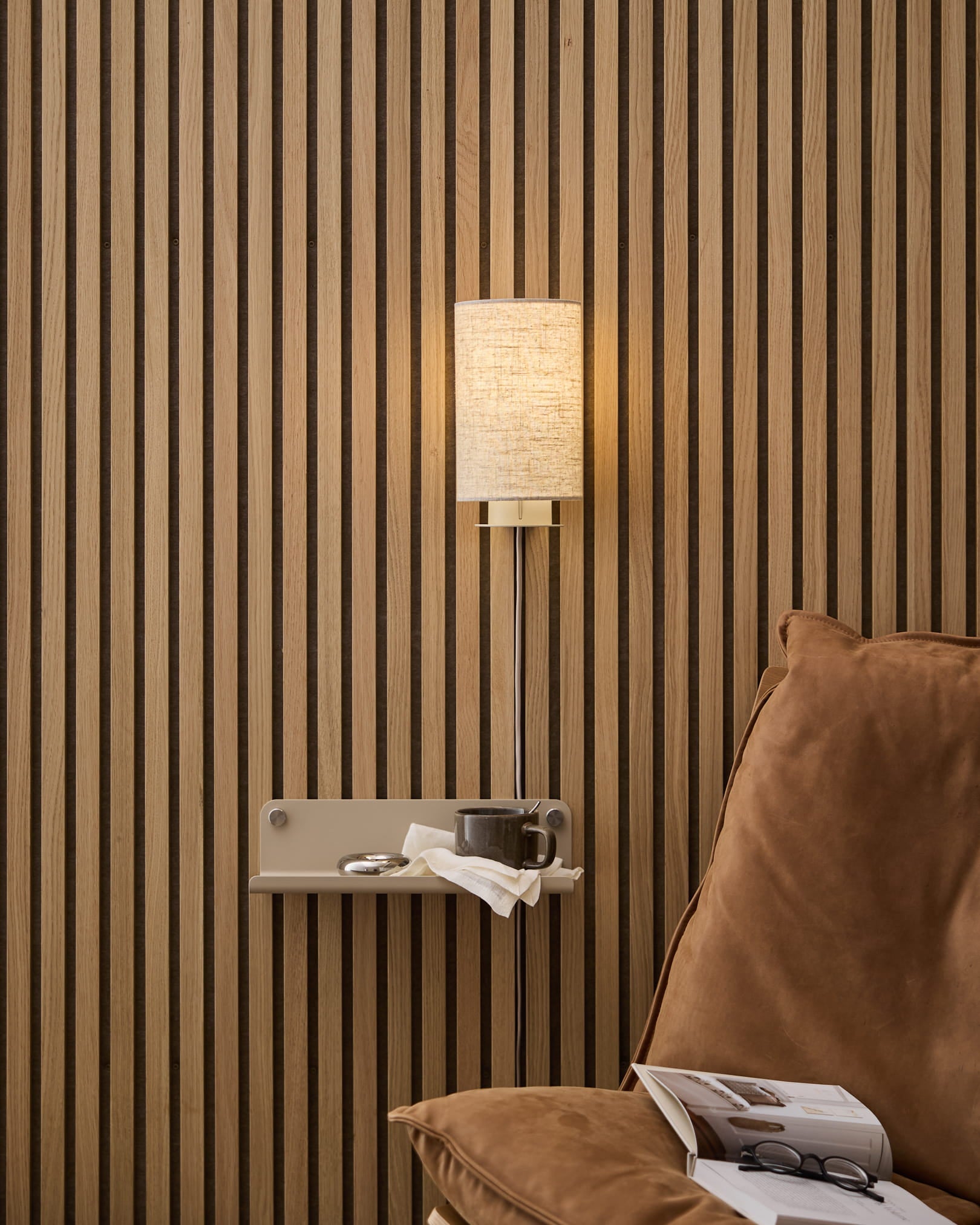 Une applique murale WoodUpp Feya en lin-coton beige, installée au-dessus d’un fauteuil cosy et d’une table d’appoint avec un livre et une tasse, sur fond de Panneau acoustique à lattes de bois nordique.