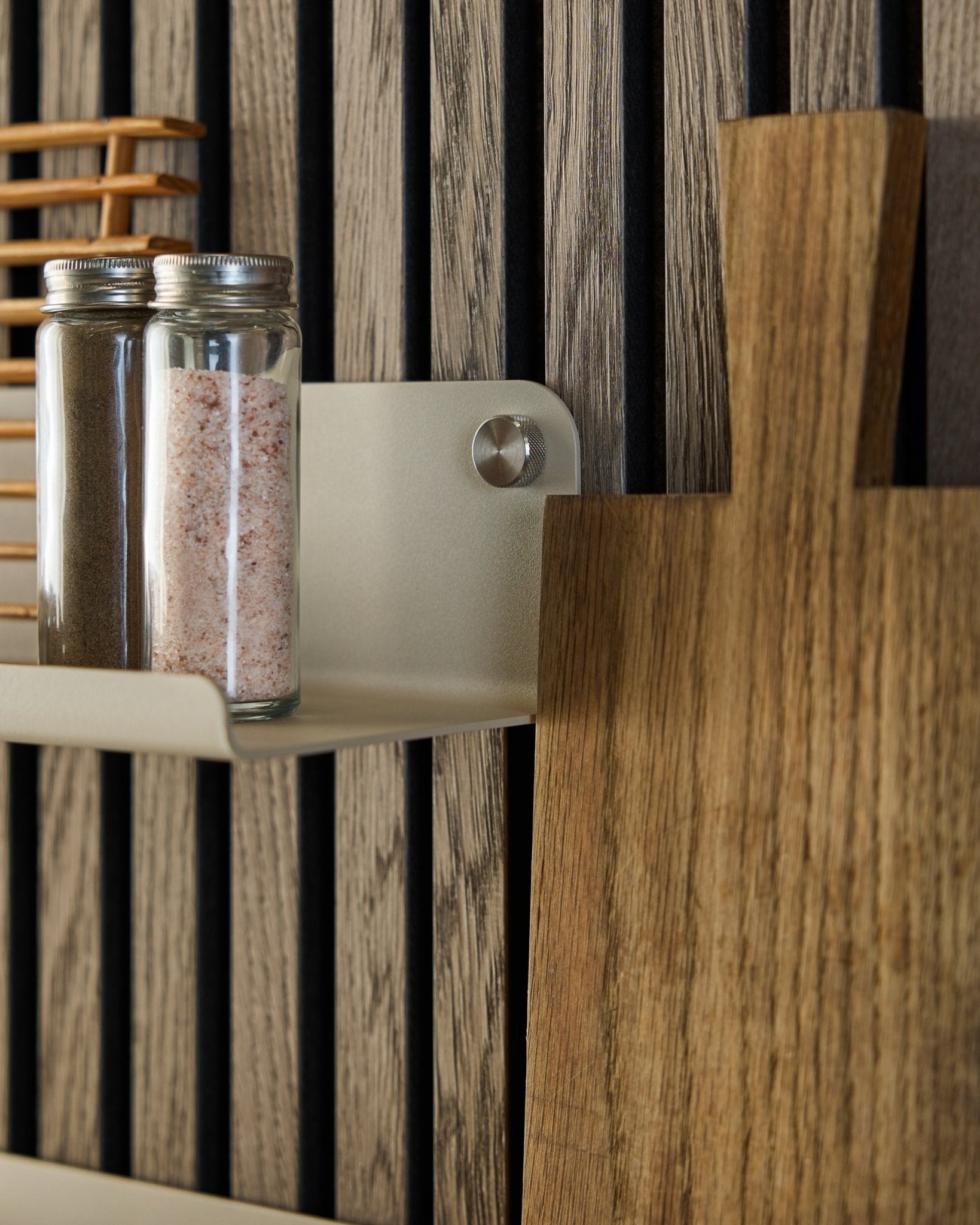 Des bocaux en verre contenant des épices et une planche à découper en bois reposent sur l’étagère large WoodUpp Norr en beige, 35,9 cm, montée sur un panneau acoustique à lattes vertical.