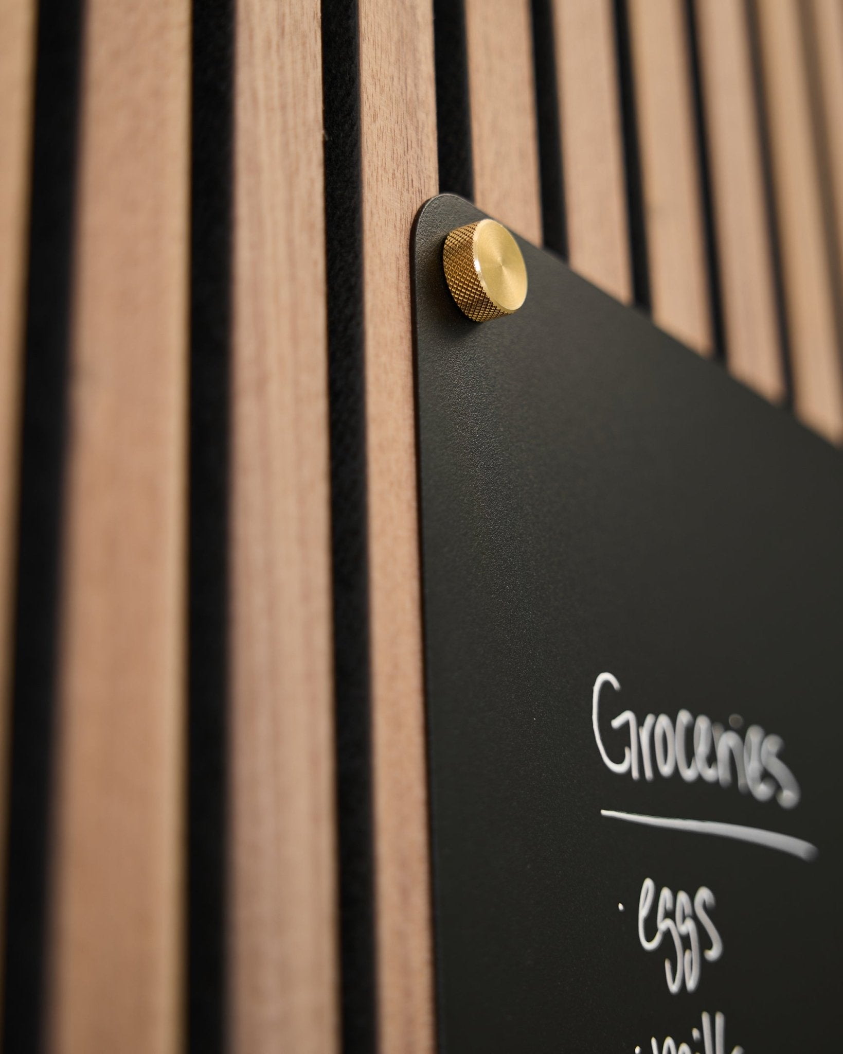 Un panneau black avec une liste de courses écrite à la main est fixé à un panneau acoustique à lattes WoodUpp à l’aide de deux vis moletées brass M5x10 mm.