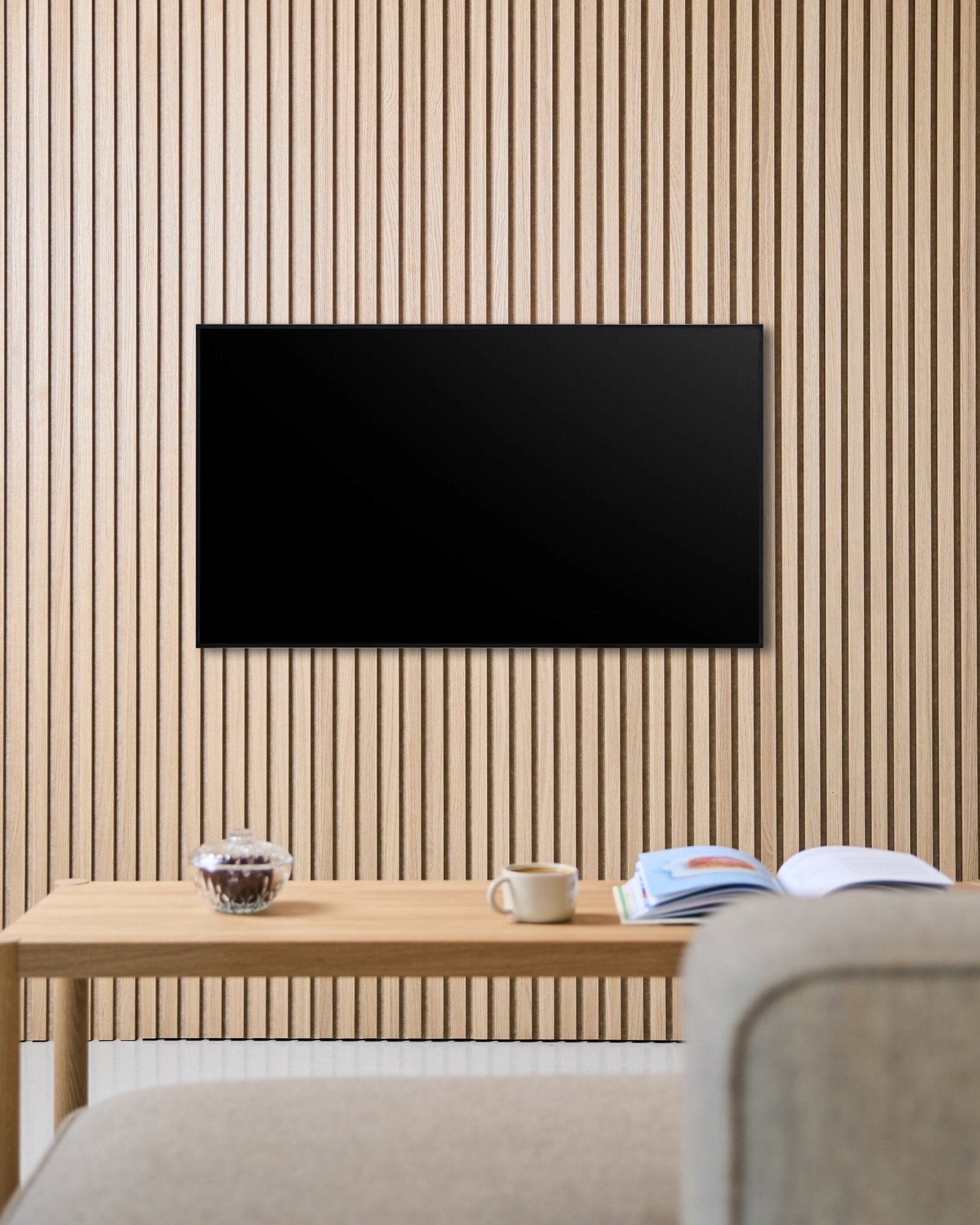 Le support TV Fjell (small) de WoodUpp fixe un écran plat sur un mur acoustique à lattes de bois Nordic Akupanel, au-dessus d’une table avec une tasse, un bol et un magazine ouvert, à côté d’un canapé beige.