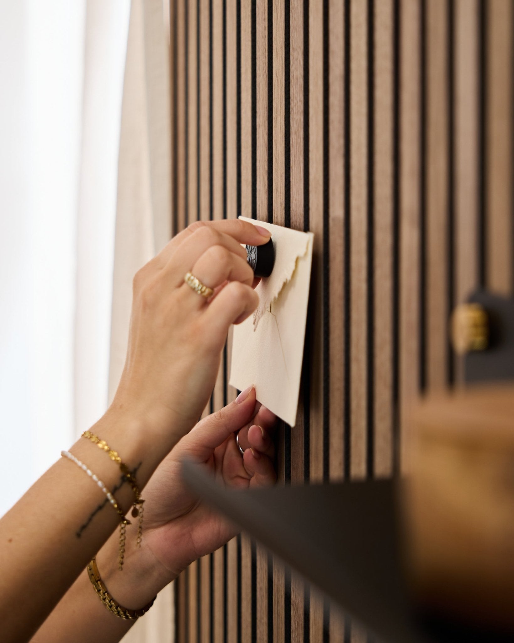 Une personne portant des bracelets et bagues dorés fixe une enveloppe sur un panneau acoustique mural à lattes de chêne WoodUpp à l’aide du set de Poster Magnet Norr en Black Oak.