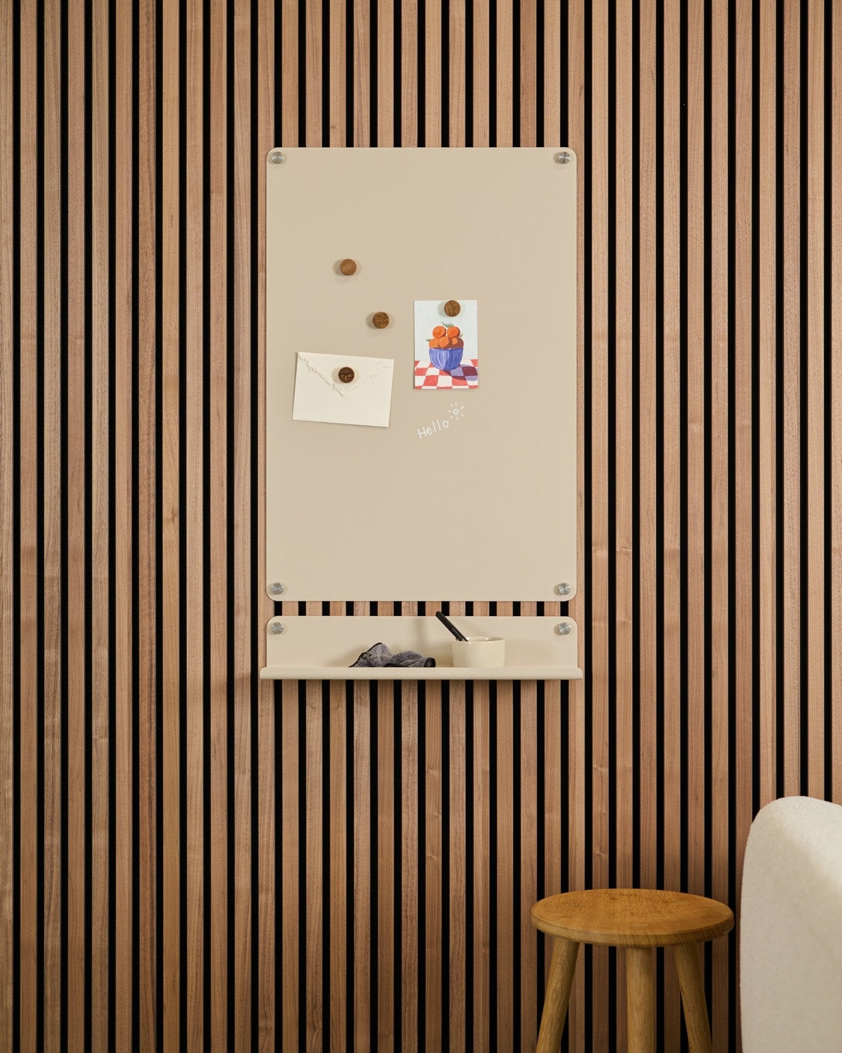 Le tableau magnétique Beige Norr de WoodUpp est fixé au-dessus d’une étagère small sur un mur en panneaux acoustiques en bois Akupanel, avec un tabouret et un canapé blanc à proximité.
