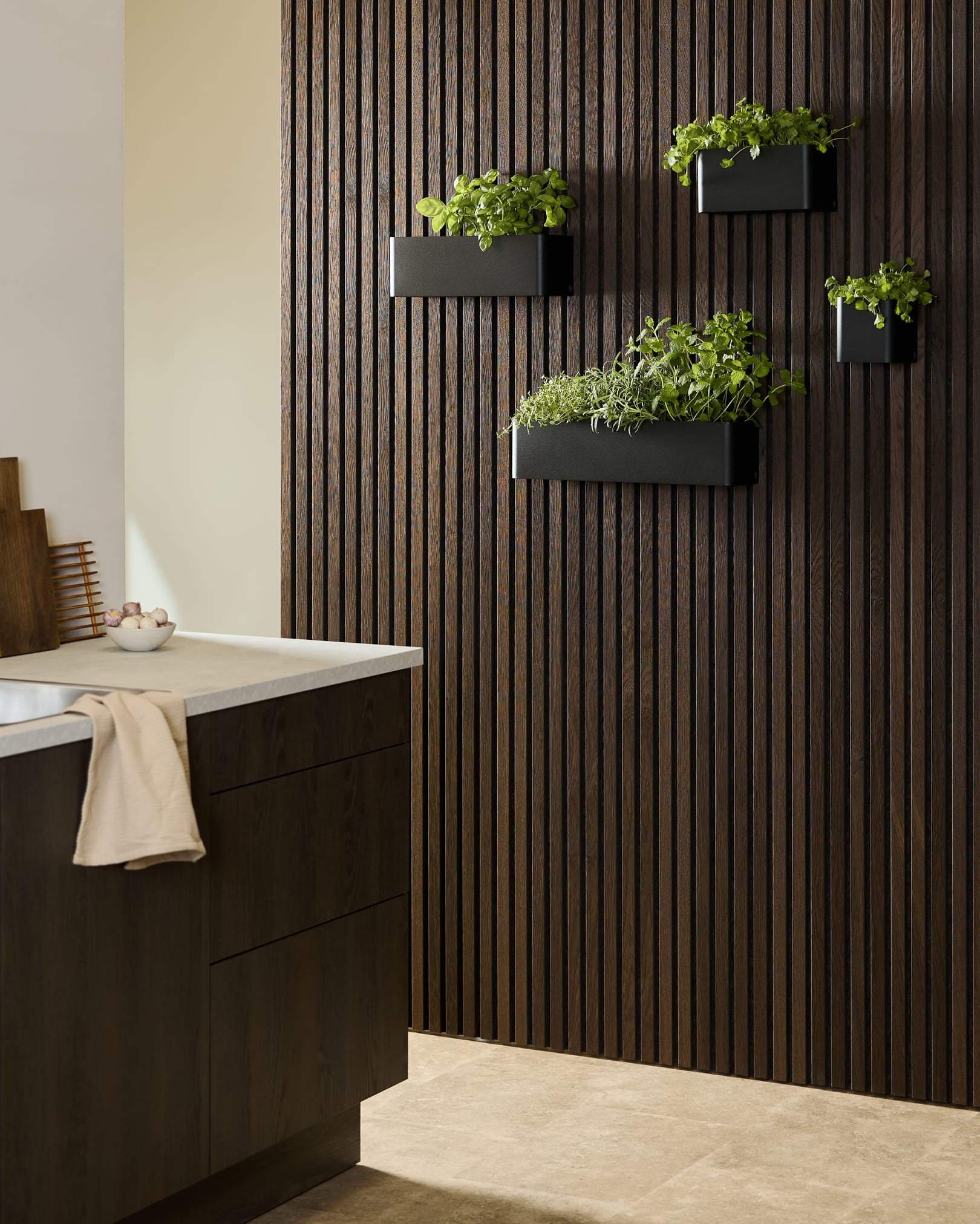 Cuisine contemporaine avec un panneau en bois foncé WoodUpp Akupanel Panneau acoustique, un Plant Box Norr black avec des herbes aromatiques, et des meubles sombres surmontés d’un plan de travail blanc.