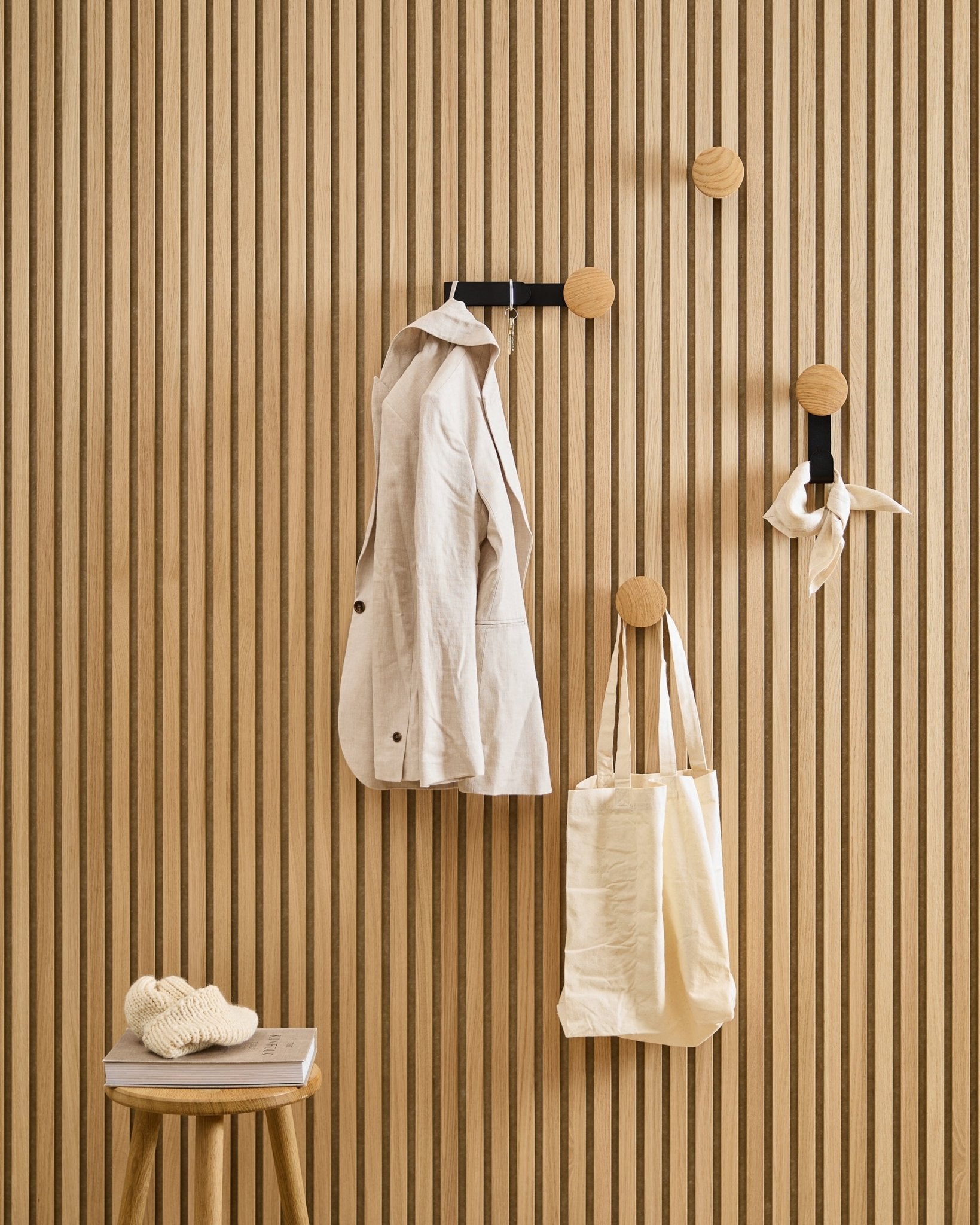 Une veste beige, un cabas et une écharpe sont suspendus au Linn Horizontal Hanger de WoodUpp en Natural Oak ; à côté, un tabouret accueille un livre et un tricot.
