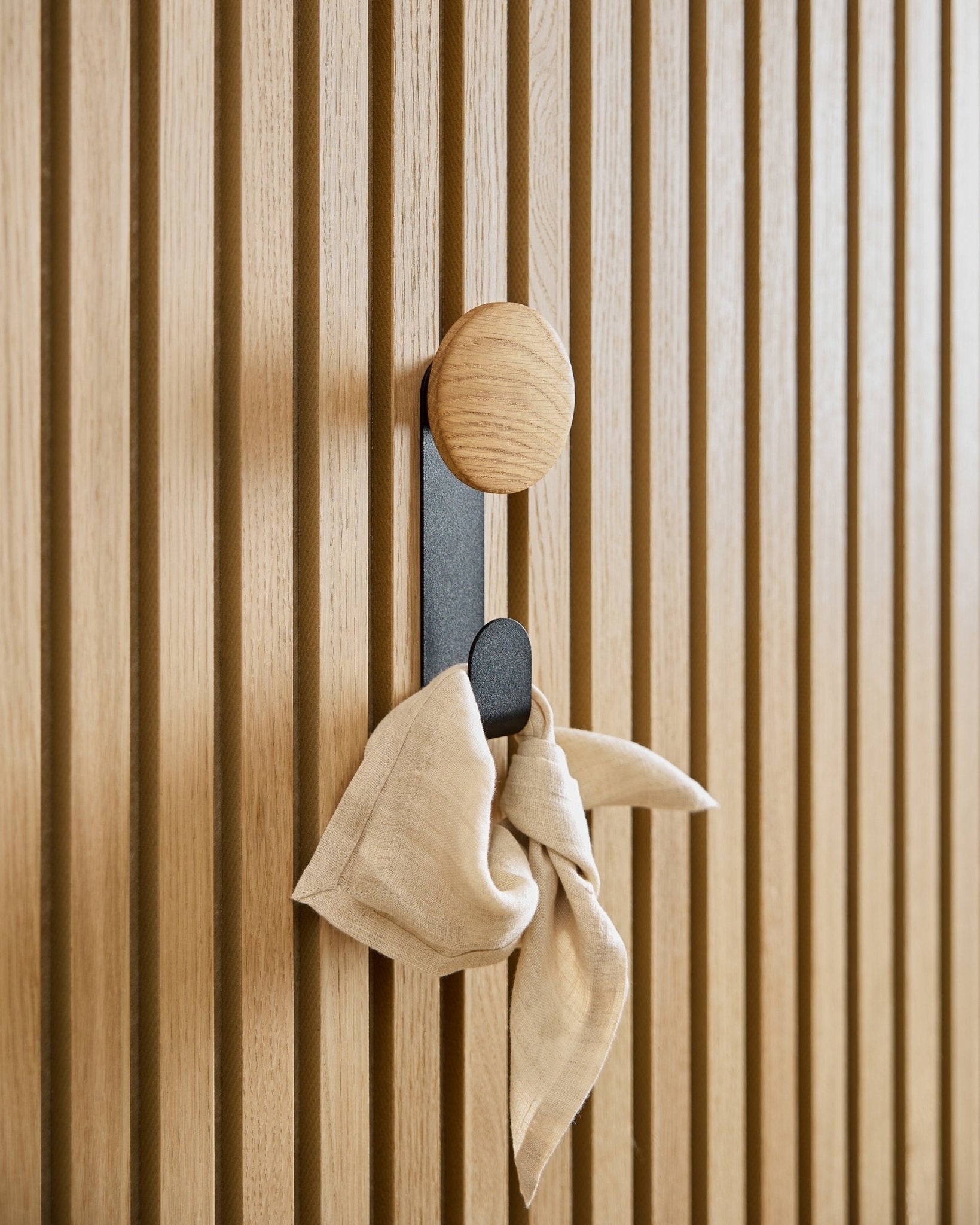 Un tissu beige est suspendu au porte-manteau Linn Vertical en Natural Oak de WoodUpp, monté sur un panneau acoustique à lattes vertical Akupanel.