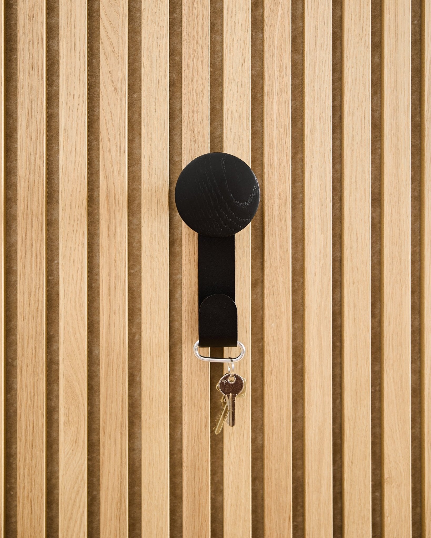 Un Hanger Linn Vertical en Black Oak par WoodUpp supporte un porte-clés avec deux clés et un médaillon rond, fixé sur un panneau acoustique en bois texturé Akupanel.