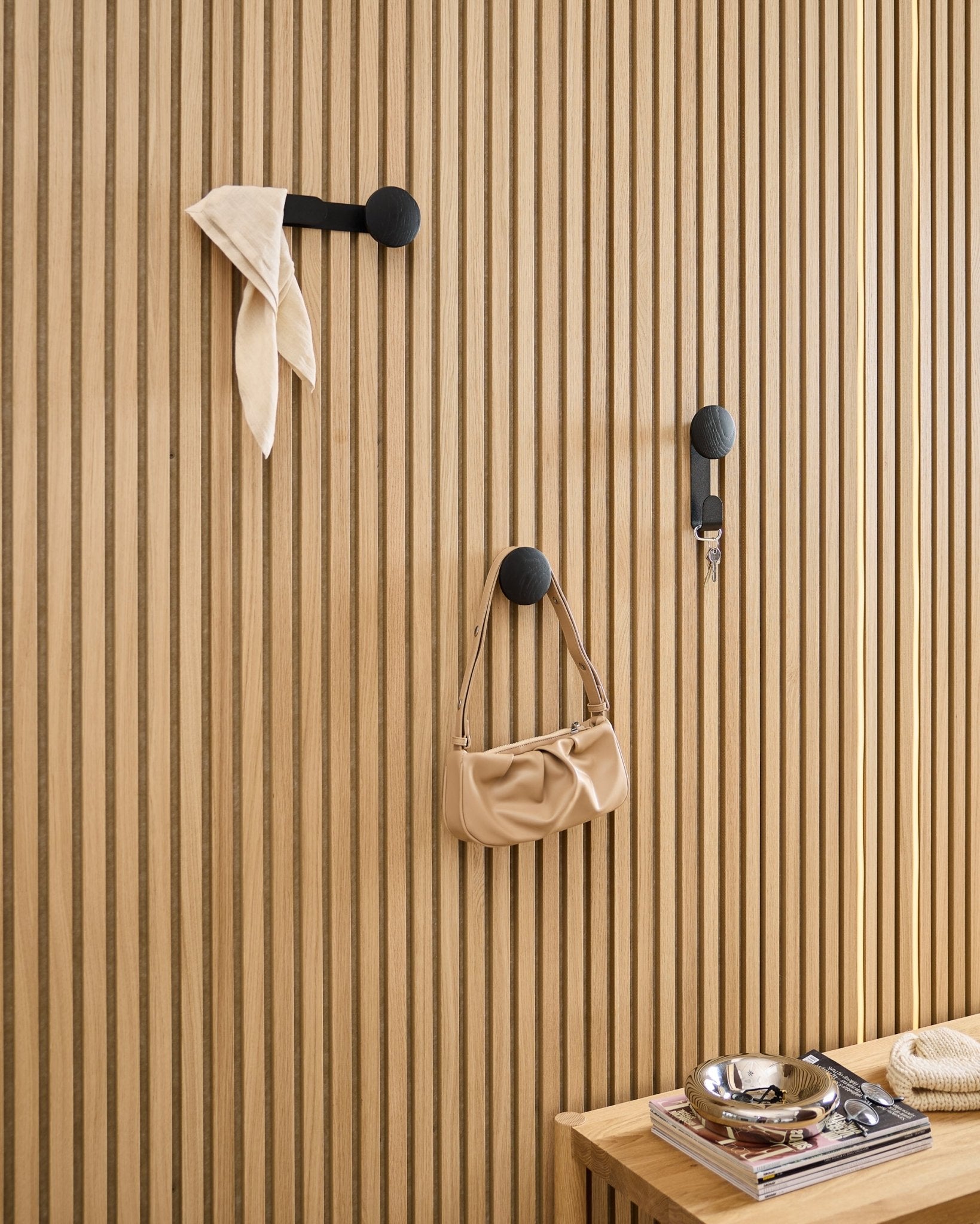 Le Linn Single Hanger WoodUpp en Black Oak accueille une écharpe, un sac beige et des clés sur un panneau acoustique en bois black Akupanel ; un banc avec des livres et une coupelle en métal se trouve à proximité.