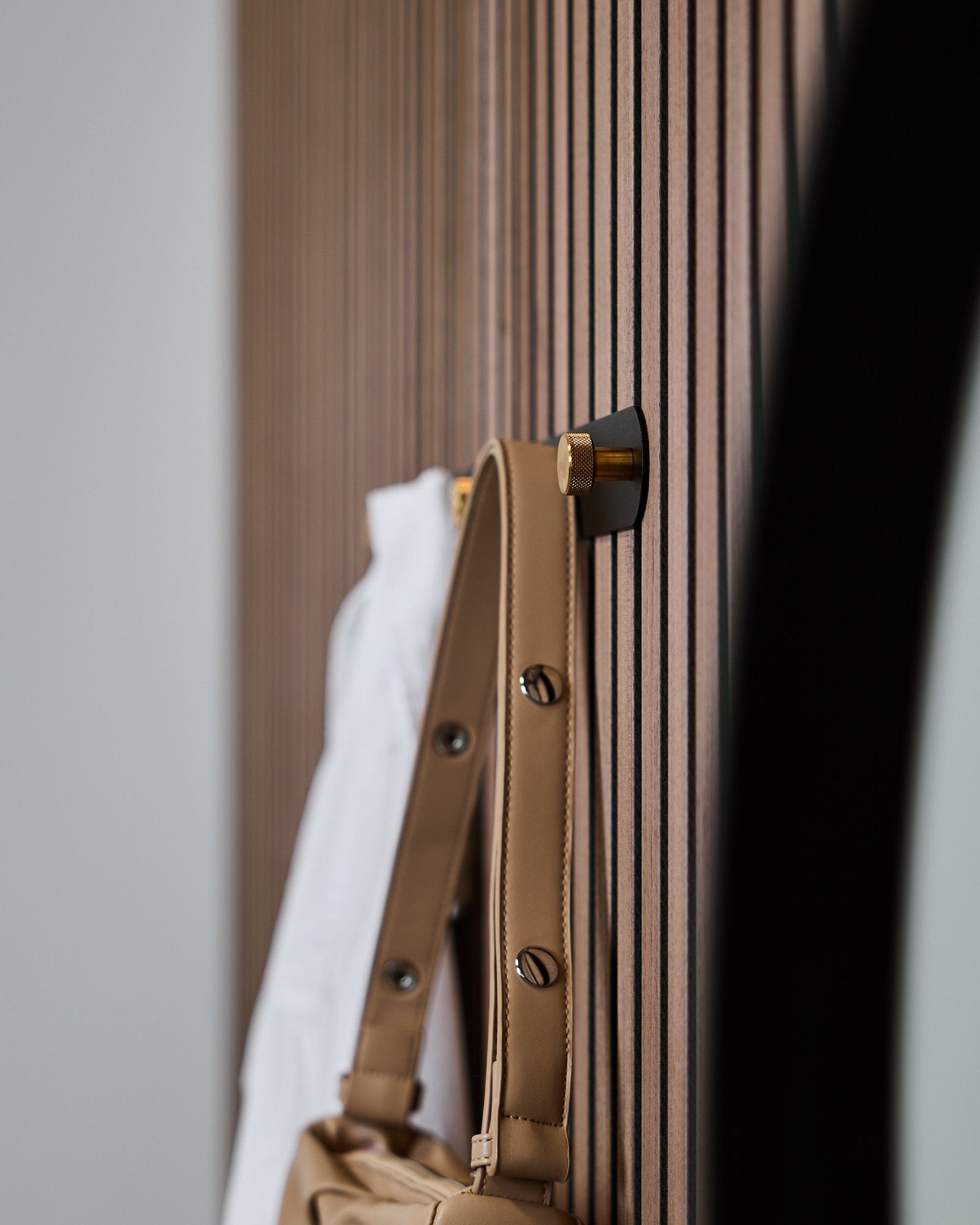 Un sac camel et une chemise blanche sont suspendus à un porte-manteau Norr Coat Hook Rack (Black/Brass, X5) de WoodUpp, monté sur un panneau acoustique à lattes vertical Akupanel.