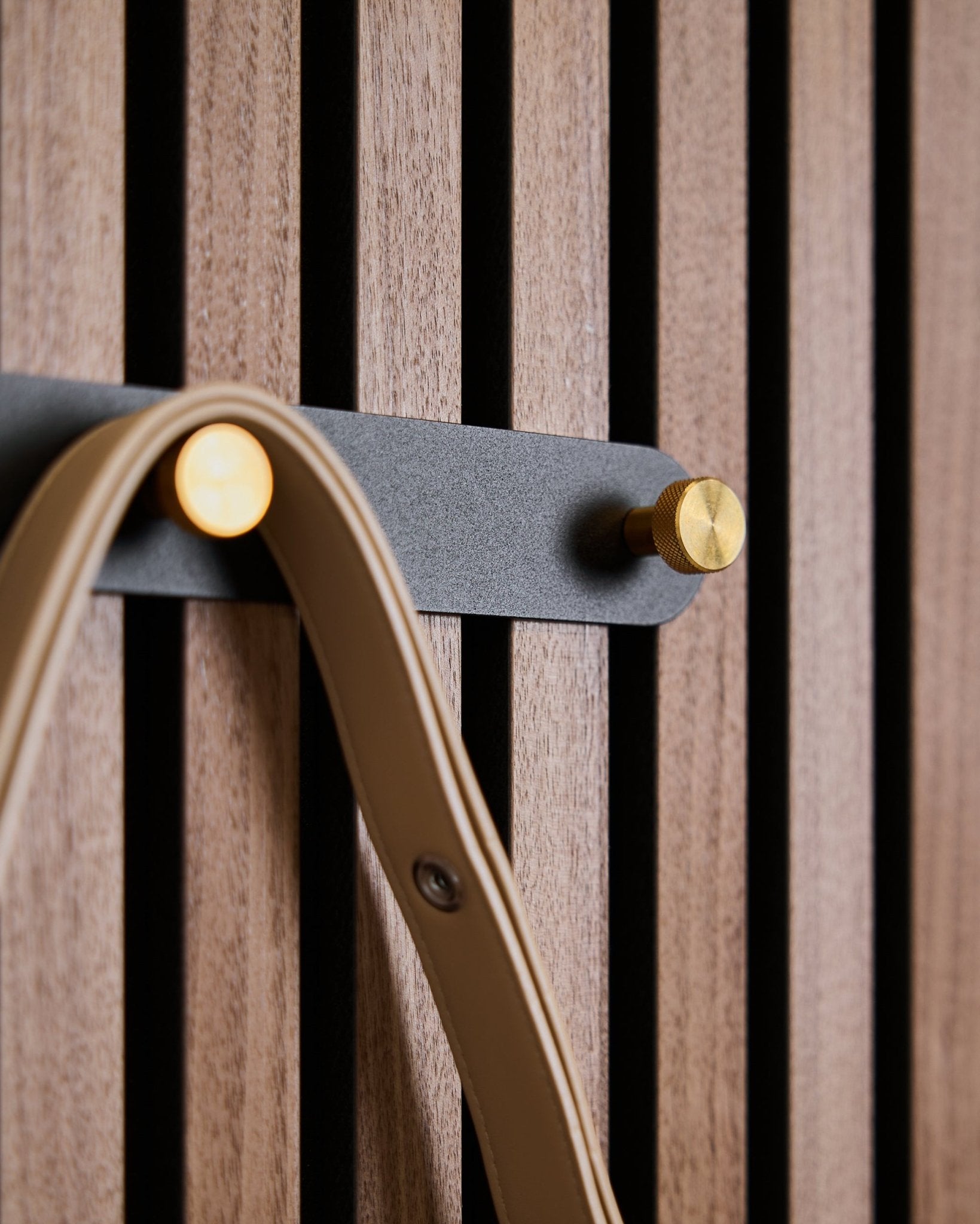 Cinq crochets black/brass du porte-manteau Norr de WoodUpp sont fixés sur un panneau acoustique à lattes en bois, chacun tenant une sangle beige.