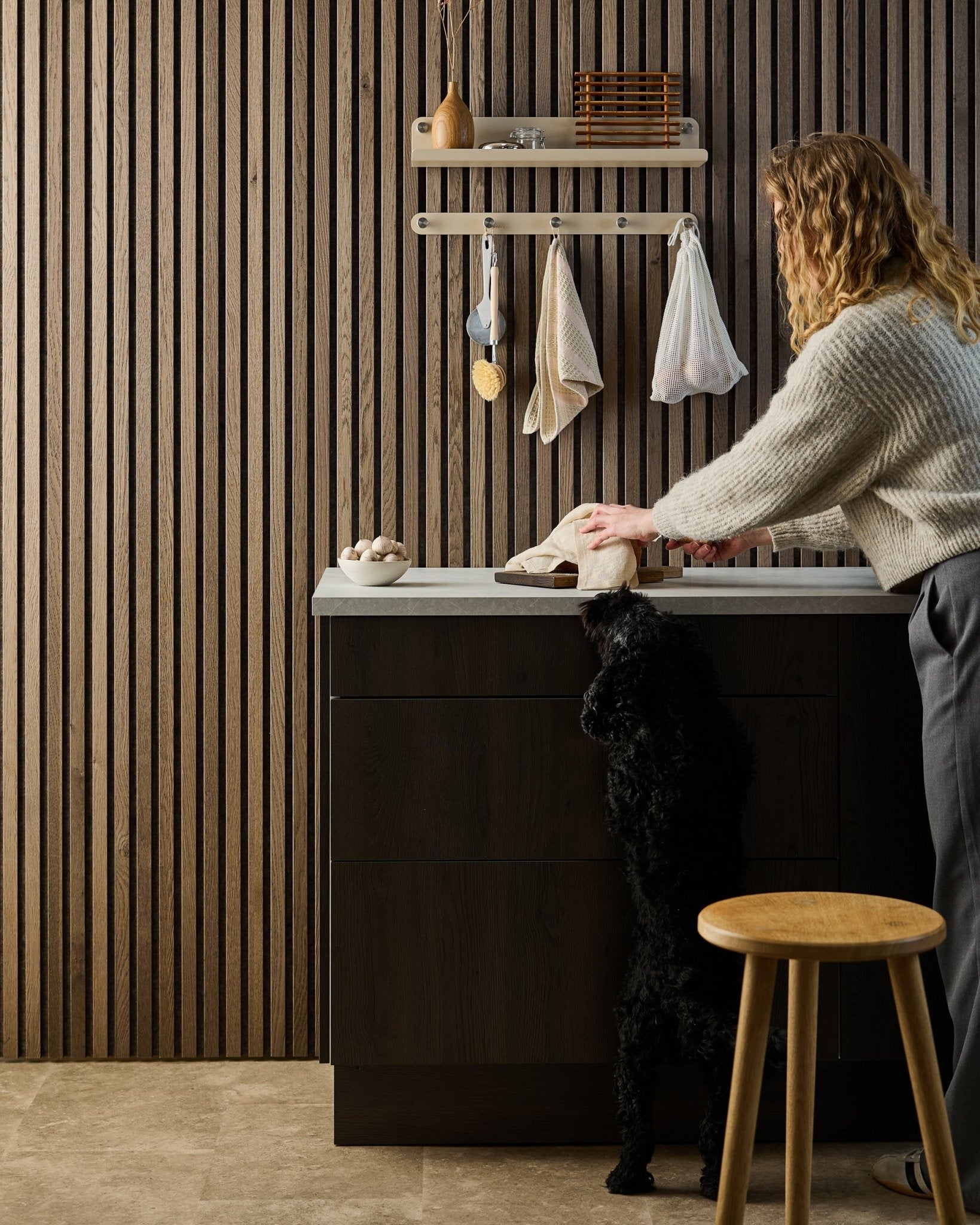 Un chien black se tient sur ses pattes arrière et essaie d’attraper de la nourriture sur un plan de travail de cuisine, avec des panneaux acoustiques en bois Nordic et des étagères en arrière-plan.