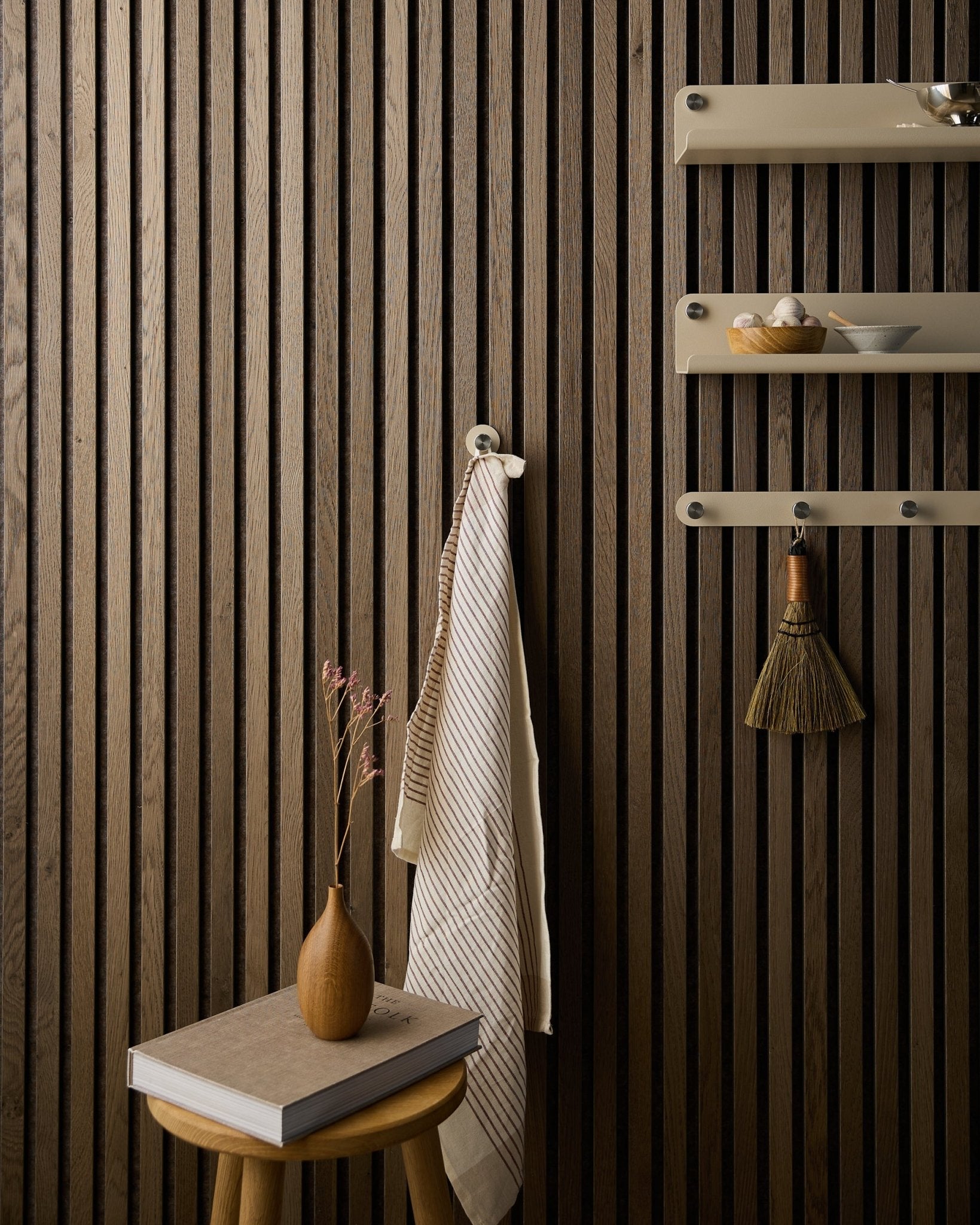 Une serviette est suspendue à une patère Norr de WoodUpp, fixée sur un mur acoustique à lattes de bois Akupanel à côté d’étagères beige, avec un balai, un tabouret, un vase et un livre.