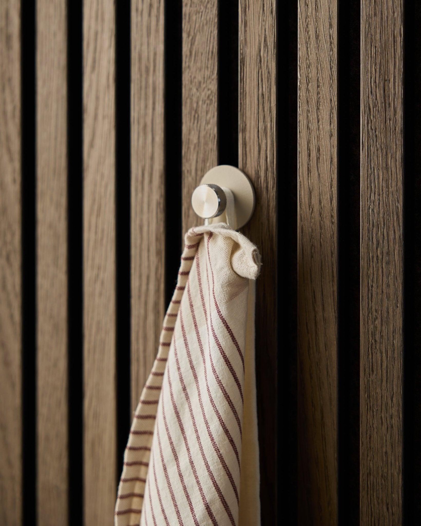 Une serviette rayée est suspendue à une patère beige Norr de WoodUpp, en acier inoxydable steel, fixée sur un panneau mural acoustique à lattes en bois texturé Akupanel.