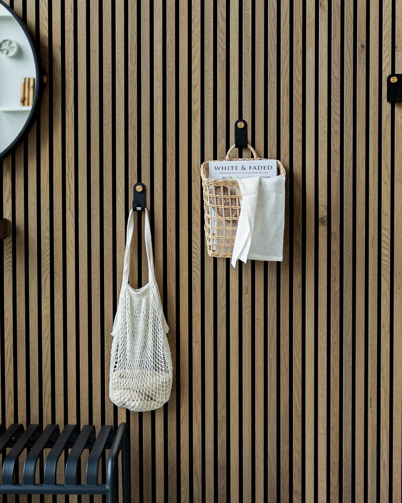 Un sac en filet et un panier sont suspendus à des patères simples Norr de WoodUpp sur un panneau acoustique en bois Akupanel Panneau acoustique Nordic ; une partie d’un miroir est visible à gauche.