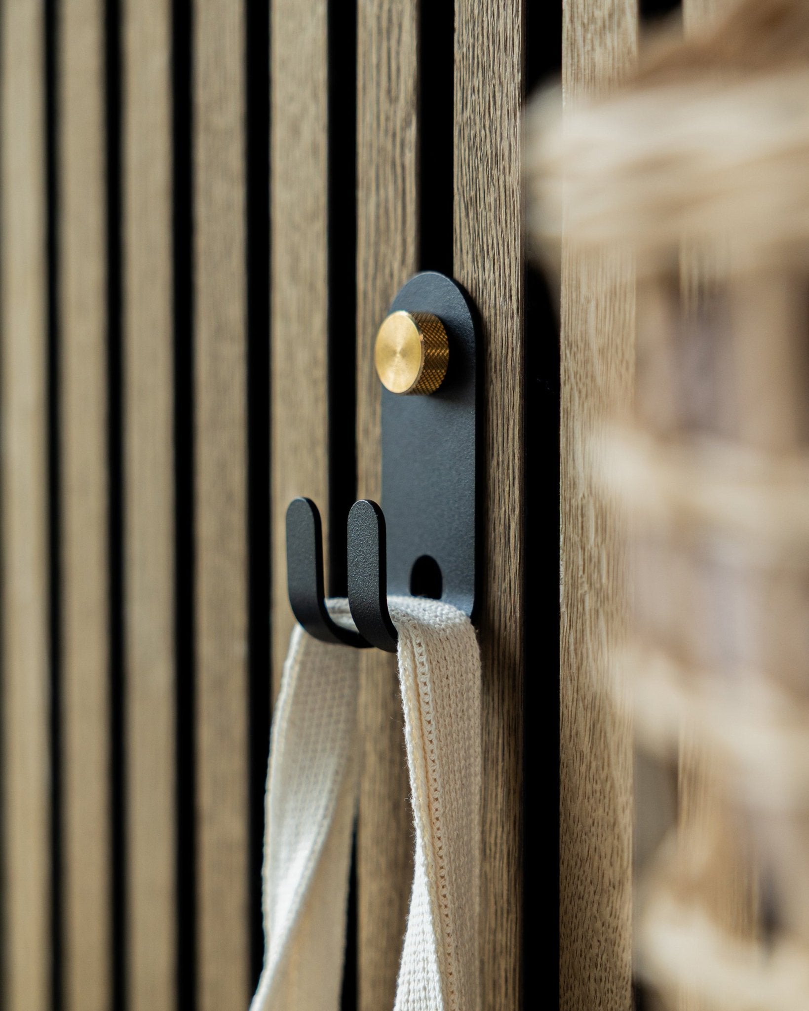 Le Norr Double Hanger de WoodUpp en black est fixé sur un panneau acoustique à lattes en bois Nordic et supporte une sangle en tissu crème-colored.