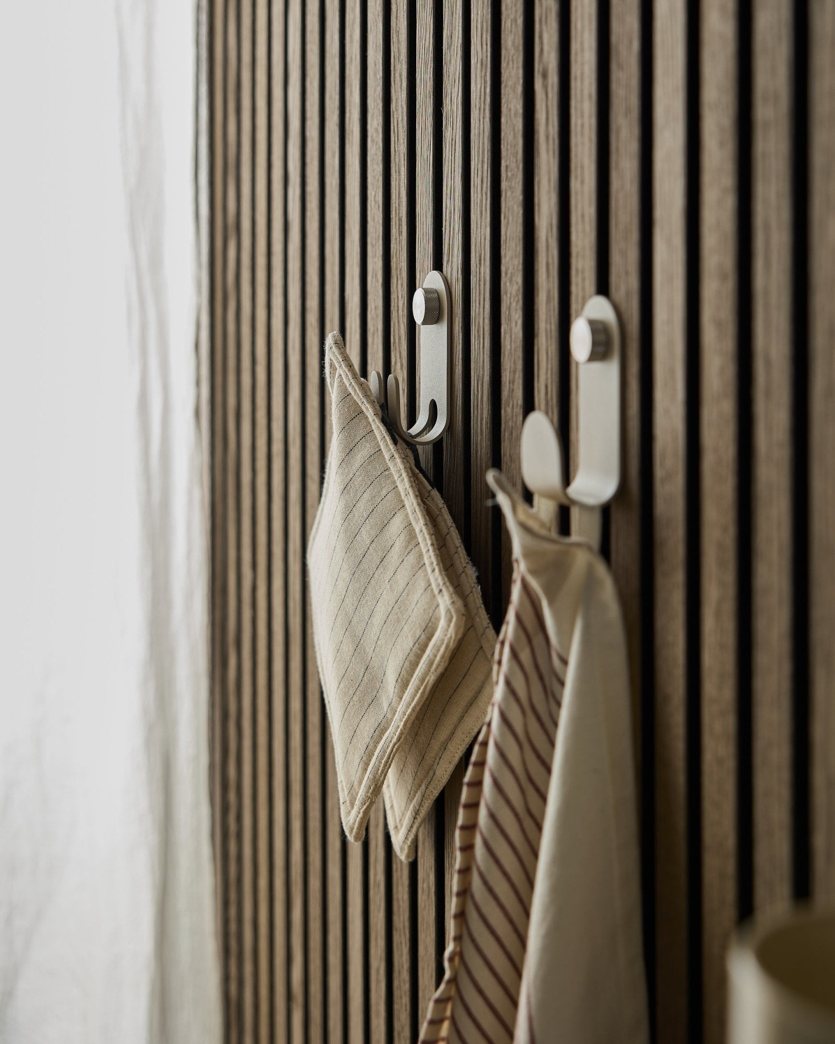 Deux torchons de cuisine rayés sont suspendus au Norr Double Hanger de WoodUpp, monté sur un panneau acoustique à lattes nordique beige Panneau acoustique, éclairé par une lumière naturelle douce.