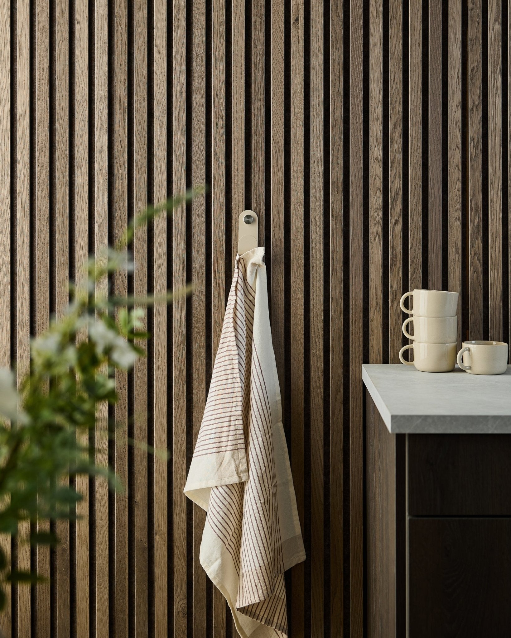 Une serviette rayée est suspendue à un beige WoodUpp Norr Single Hanger fixé sur un panneau mural à lattes en bois Panneau acoustique ; des tasses empilées et une plante sont posées sur le plan de travail en dessous.