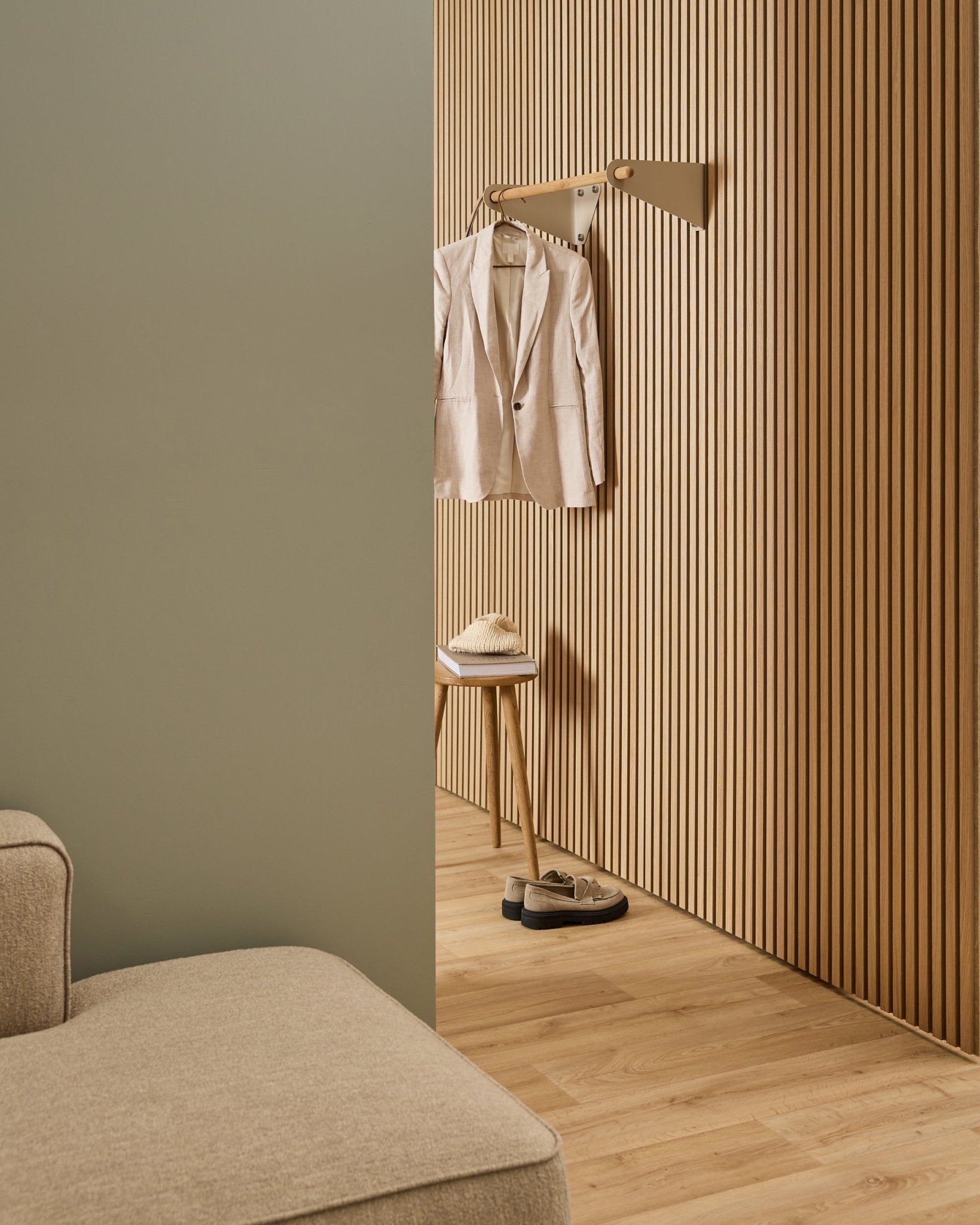 Pièce minimaliste avec canapé beige, mur acoustique à lattes Akupanel, portemanteau WoodUpp Linn en beige (75,9 cm), tabouret avec chapeau et chaussures sur un sol en bois clair.