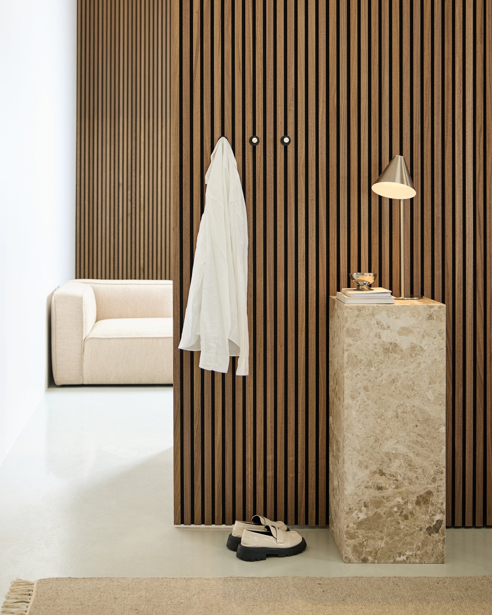 Pièce minimaliste avec un mur acoustique nordique à lattes de bois Akupanel, une chemise blanche suspendue à des patères murales, un piédestal en pierre avec une lampe et des livres, un canapé beige et des chaussures blanches au sol.