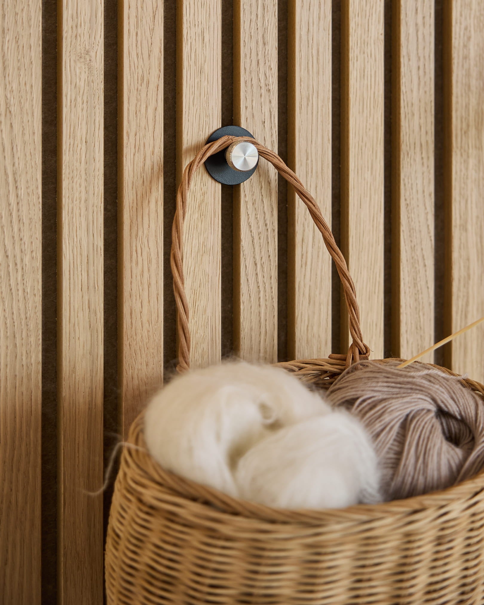Un panier en osier blanc et en fil beige est suspendu à une patère Norr Coat Hook / X1, Black/Stainless Steel de WoodUpp, montée sur un panneau acoustique à lattes Akupanel.