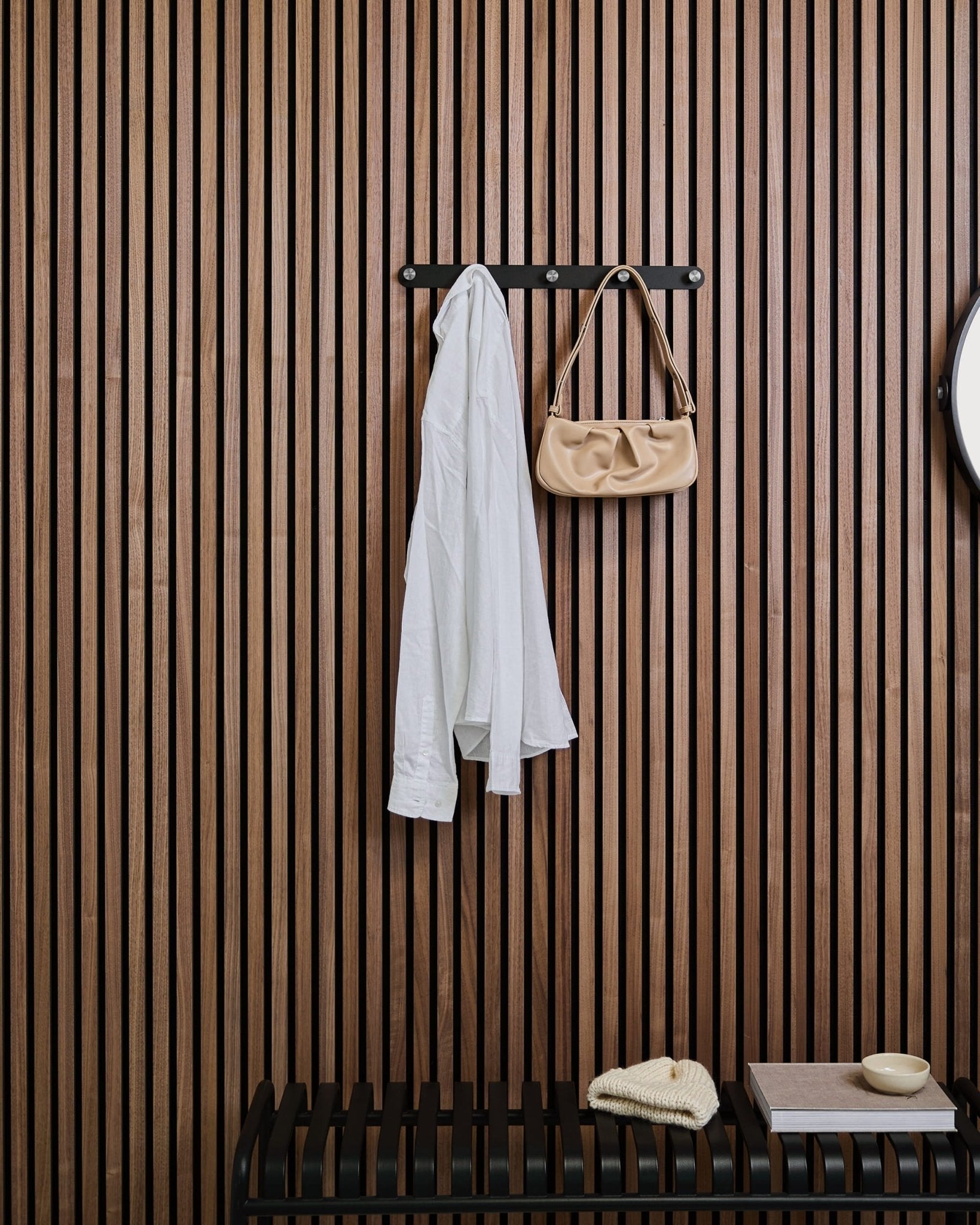 Une chemise blanche et un sac à main beige sont suspendus à la patère murale Norr de WoodUpp au-dessus d’un banc, devant un panneau mural acoustique à lattes en bois Akupanel avec quelques objets déco.