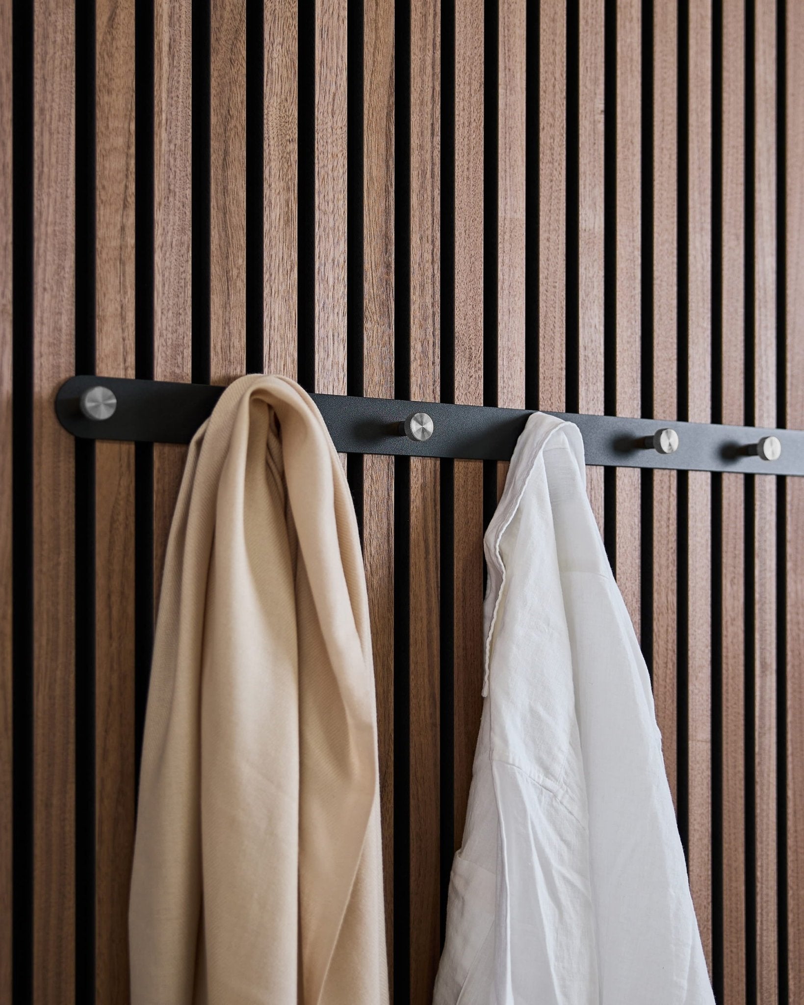 Une écharpe beige et une chemise blanche sont suspendues au porte-manteau à patères WoodUpp Norr X7 Black/Stainless Steel, fixé sur un panneau acoustique à lattes nordique.