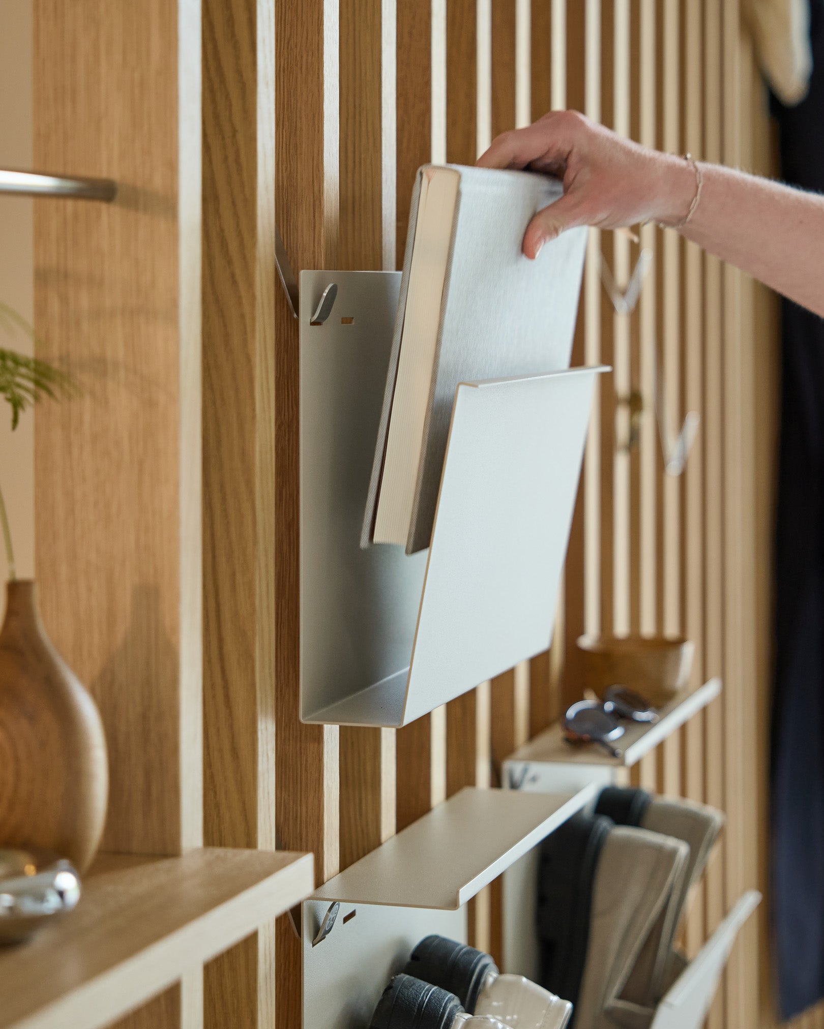 Une main pose Nell | Magazine Storage / Beige de WoodUpp sur une étagère en steel thermolaqué montée sur un panneau acoustique nordique en bois Panneau acoustique, avec d’autres objets à proximité.