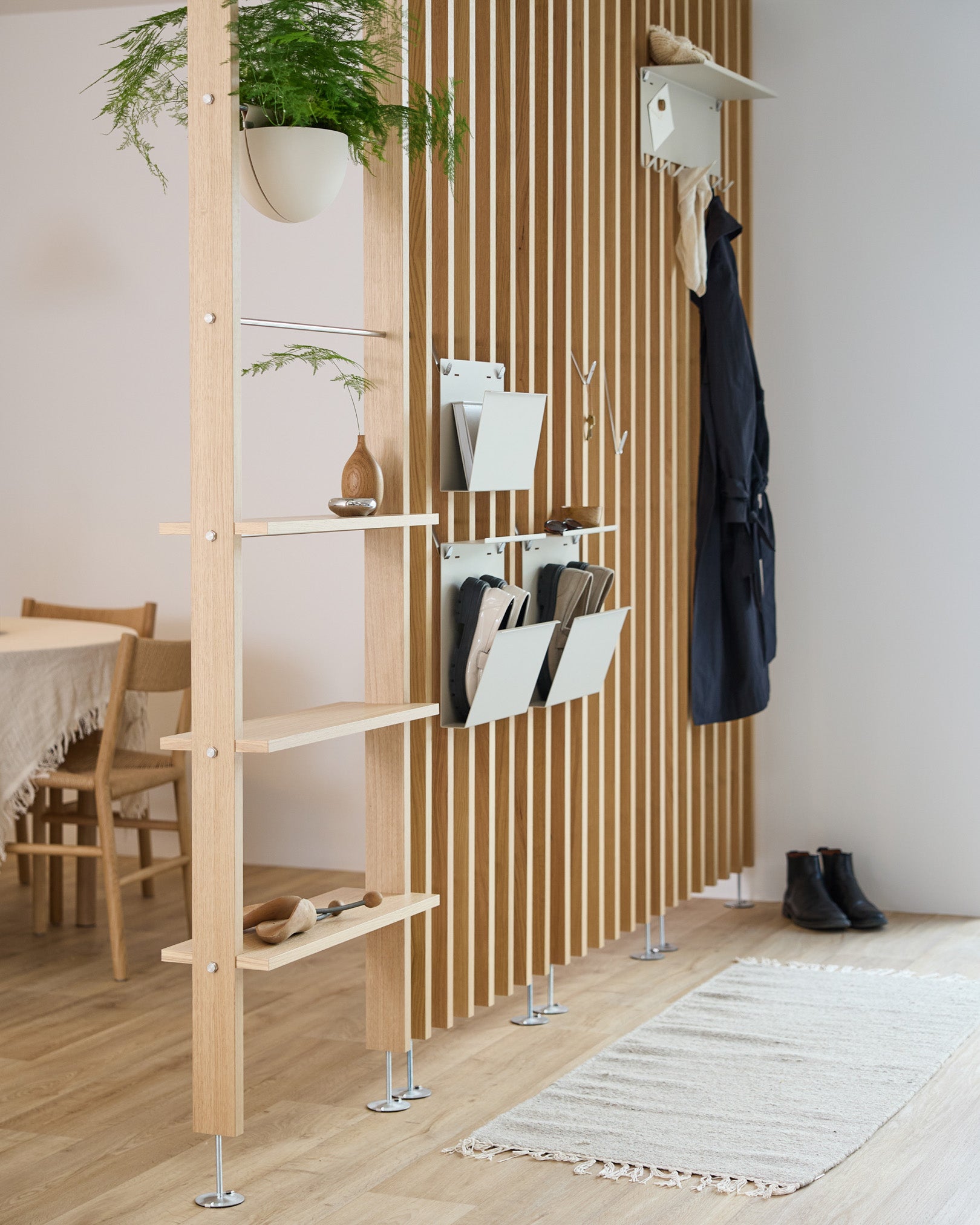 Le porte-revues Nell de WoodUpp en beige est intégré à un séparateur de pièce avec des étagères en steel, des bacs de rangement et des patères ; on aperçoit derrière un coin repas et un tapis.