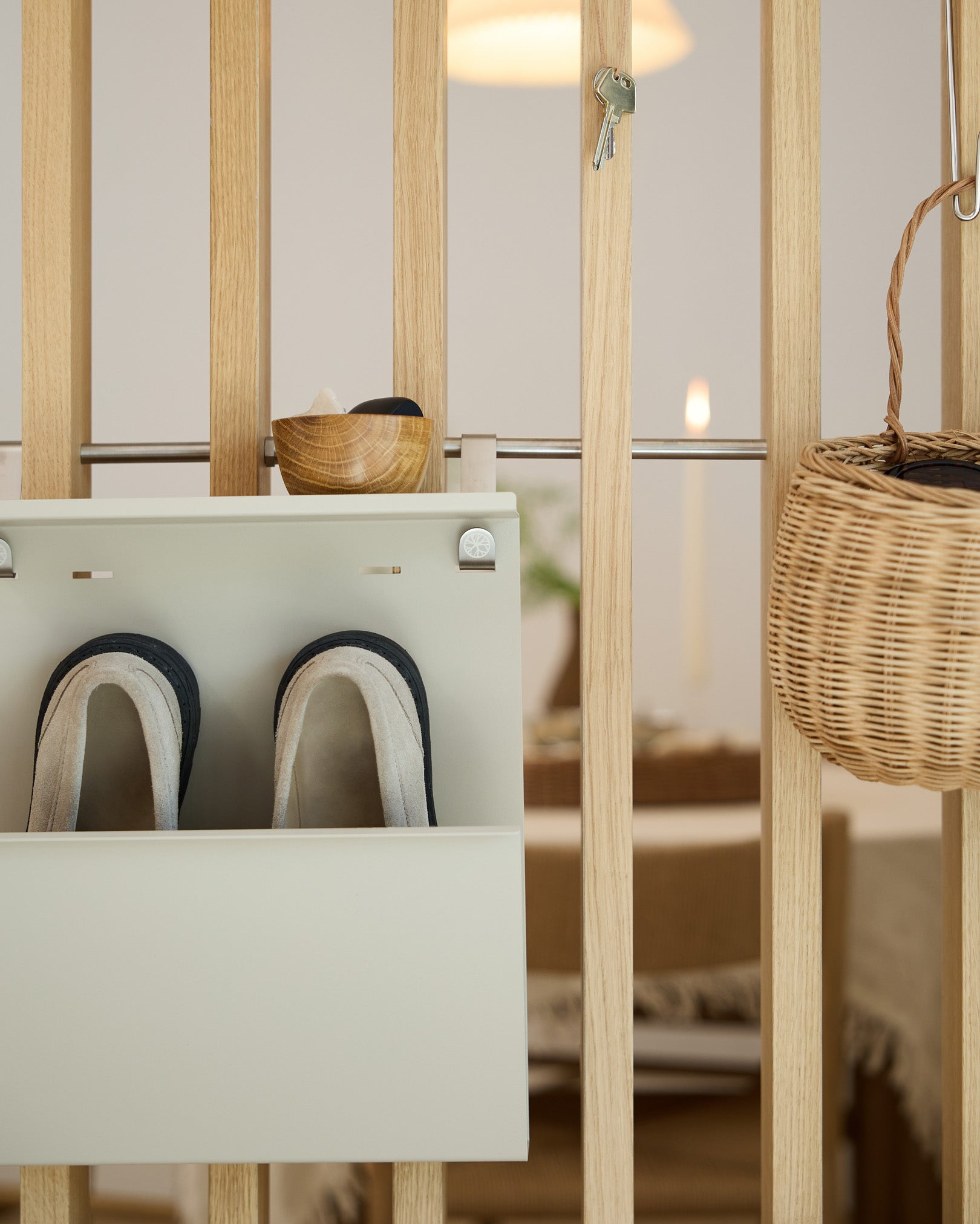 Le rangement à chaussures Nell Beige de WoodUpp présente des chaussons, un bol et un panier en osier sur un séparateur de pièce en bois ; l’espace repas est visible à l’arrière-plan.