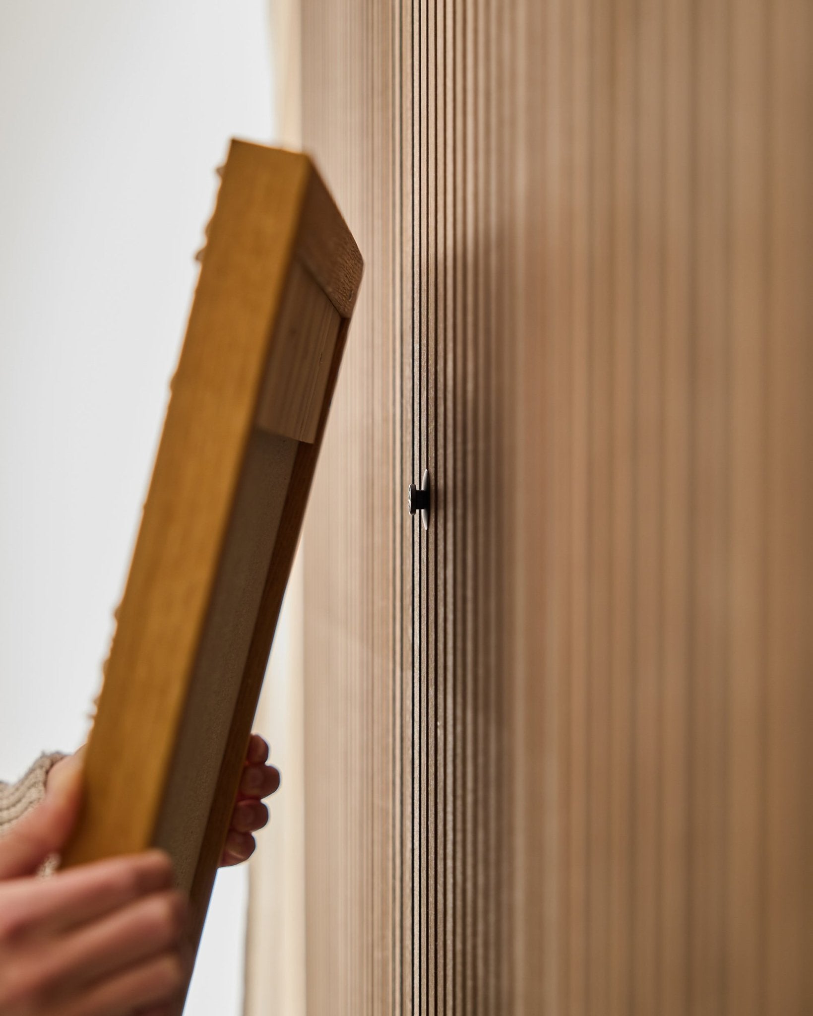 Une personne utilise le kit WoodUpp Dupps | Original pour monter un cadre de panneau acoustique en bois à lattes Akupanel, en se concentrant sur la fixation d’un crochet small black près des lattes vertical.