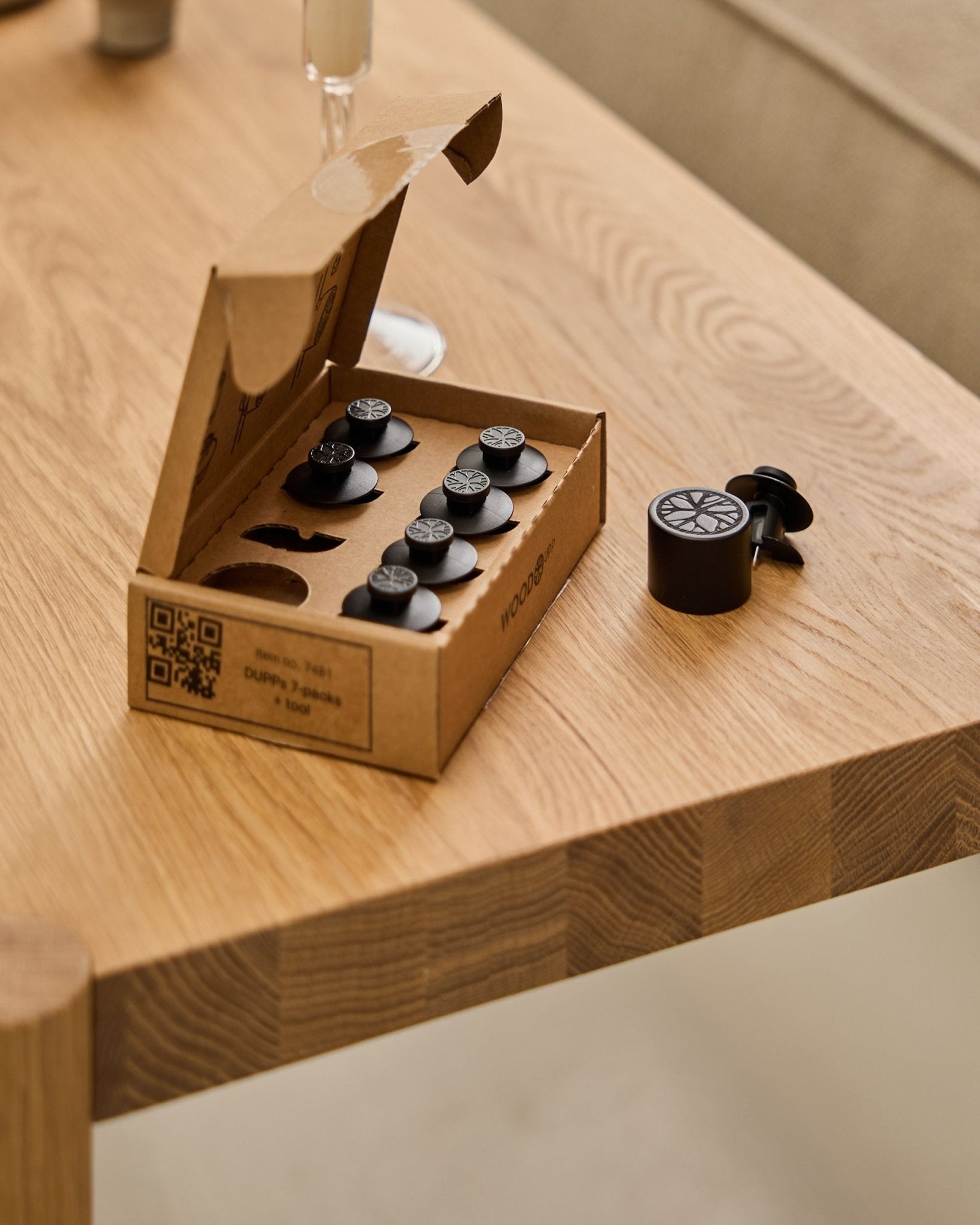 Le set WoodUpp Dupps Original (7 pièces et 1 outil) disposé sur une table en bois, avec une pièce Dupps et une partie d’un verre visibles sur le bord.