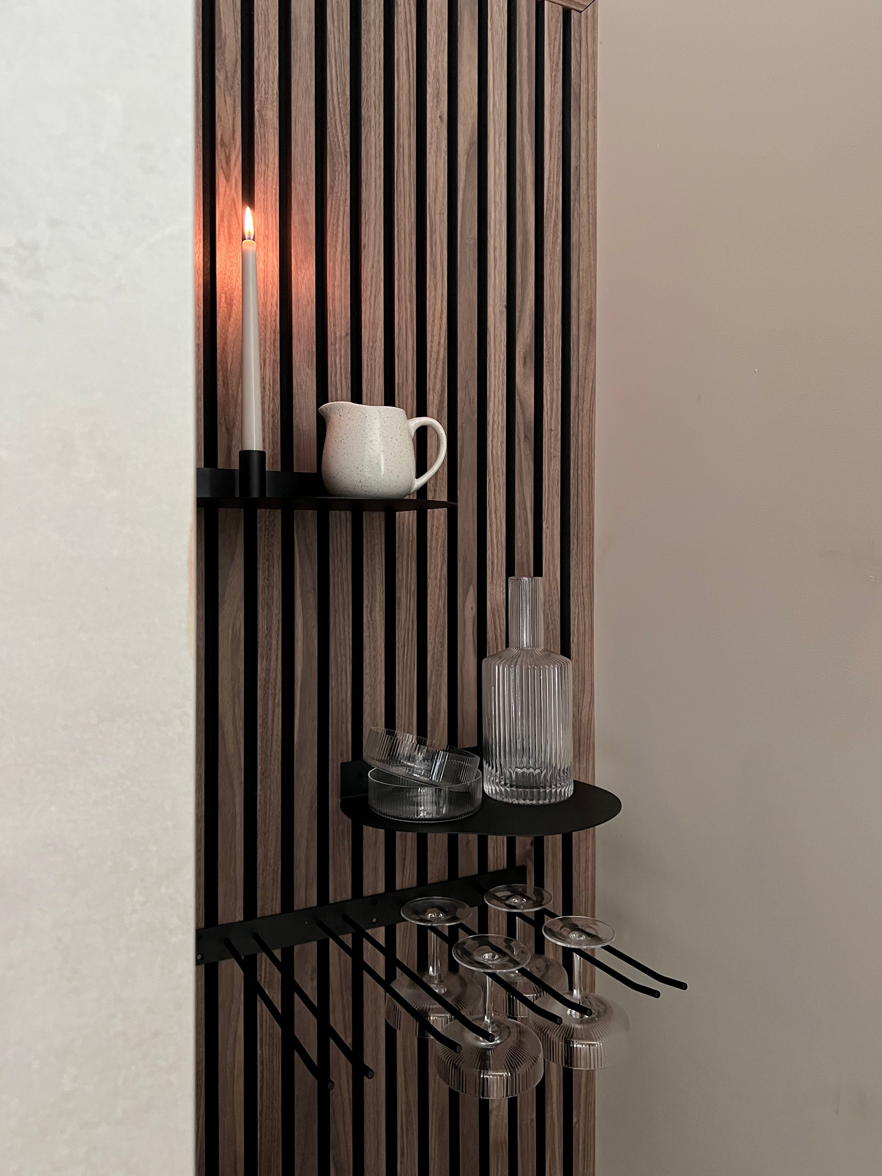 Panneau acoustique nordique moderne avec des étagères noires contenant une bougie allumée, un pichet blanc, une bouteille en verre et des verres à vin à pied suspendus en dessous.