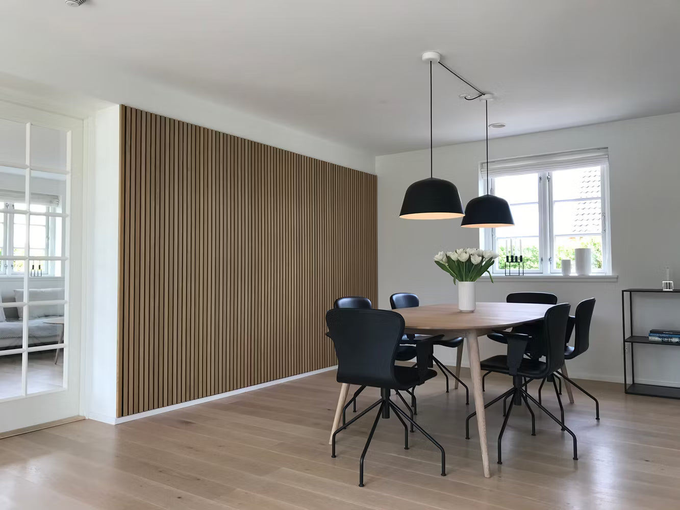 Salle à manger moderne avec sol en bois, accent acoustique en Akupanel, chaises noires, table en bois, lampes suspendues et centre de table floral blanc.