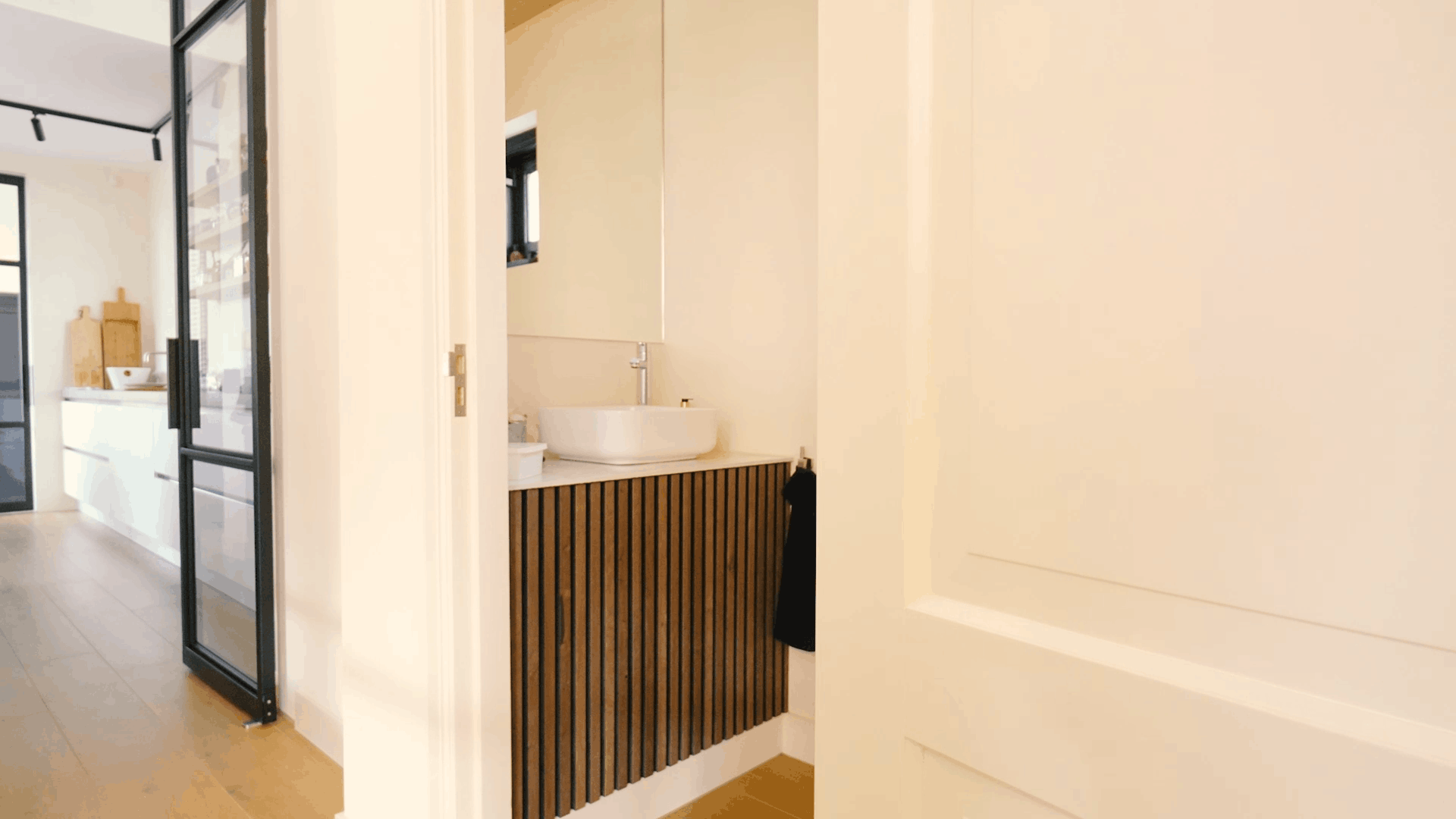 Salle de bains moderne avec lavabo rond, meuble-lavabo acoustique en bois, grand miroir et porte blanche à côté dun couloir lumineux et dune cuisine contemporaine.